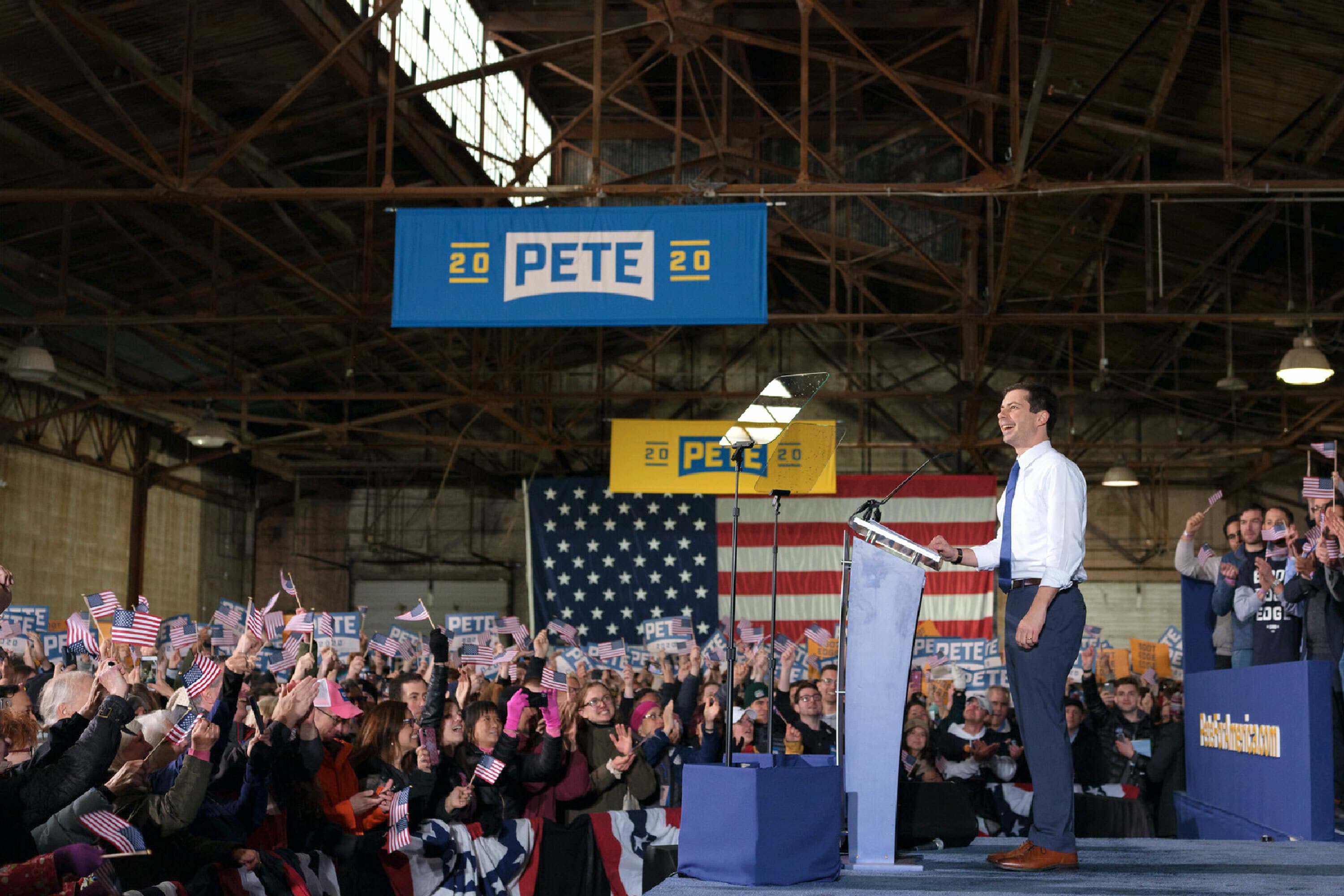 A man stands at a podium on a stage, speaking to a crowd holding small American flags and signs with "PETE" on them. Behind the speaker, an American flag and "PETE 2020" signs are displayed. The location appears to be an indoor event space with exposed beams.