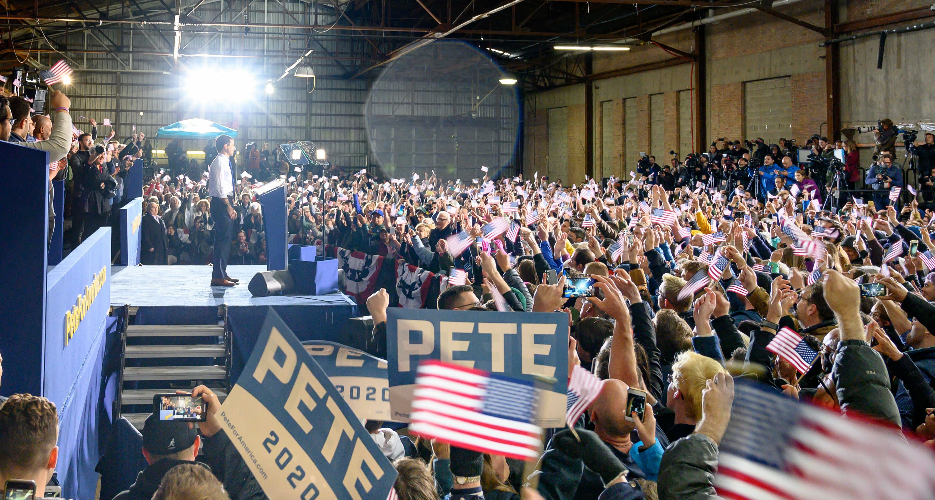 A speaker stands on a stage addressing a large, enthusiastic crowd in a warehouse. Attendees hold up American flags and signs that read "PETE 2020." Several media cameras are set up, recording the event. The atmosphere is lively with patriotic symbols prominently displayed.