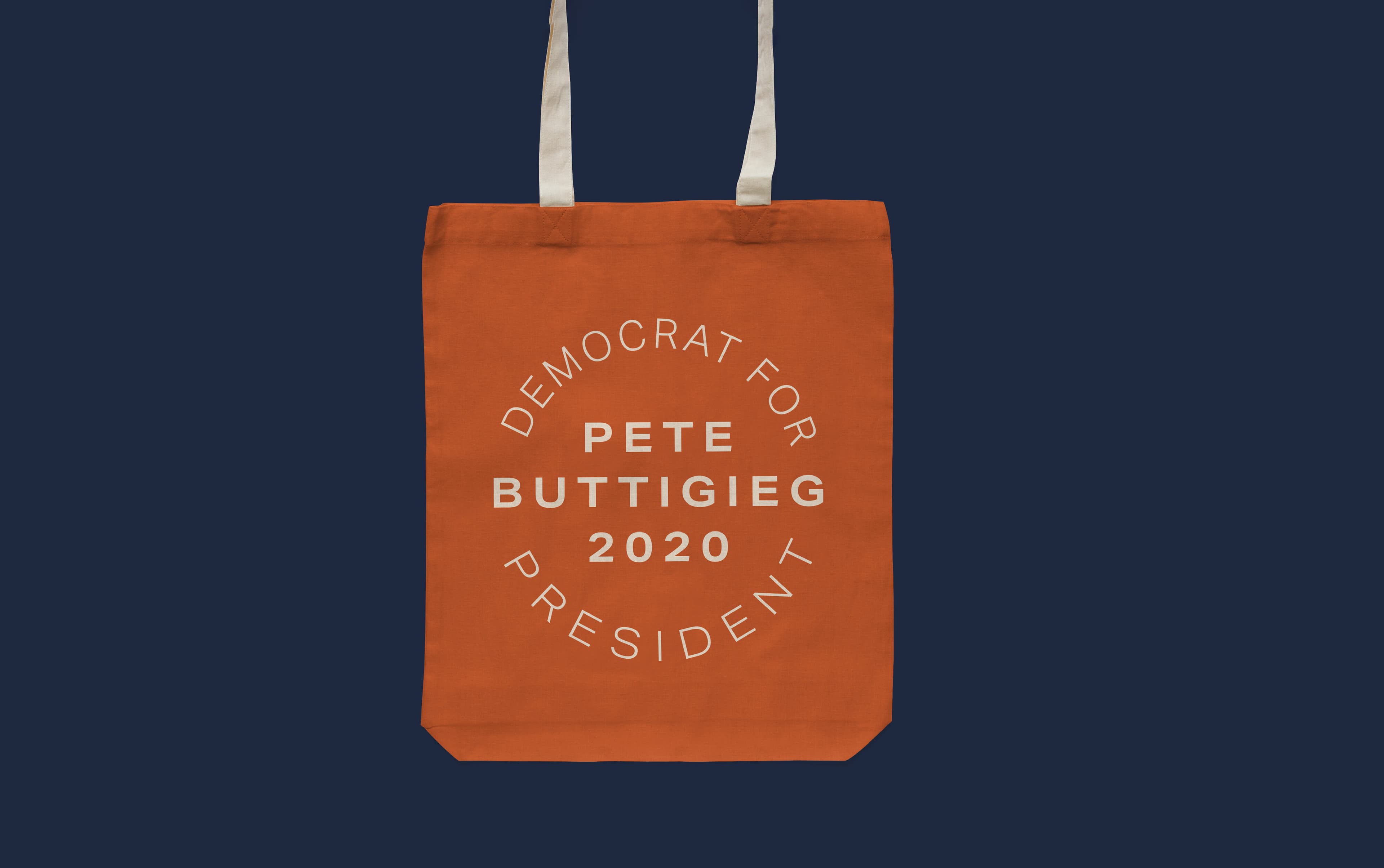 A bright orange tote bag against a dark blue background has the words "DEMOCRAT FOR PRESIDENT PETE BUTTIGIEG 2020" printed in white text, arranged in a circular pattern in the center of the bag. The bag features white handles.
