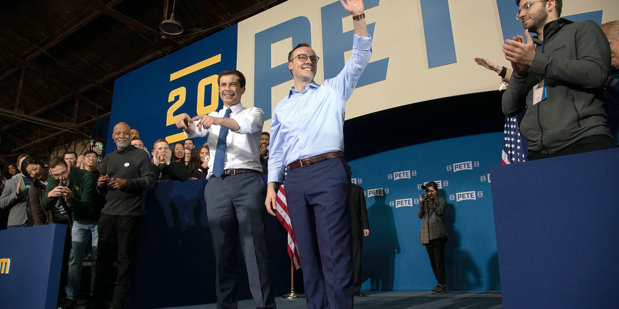 Two people standing on a stage wave to a crowd. The person on the right holds an American flag. Behind them is a large sign with the name "PETE" and the year "2020." Several people on the stage and in the audience clap and smile.