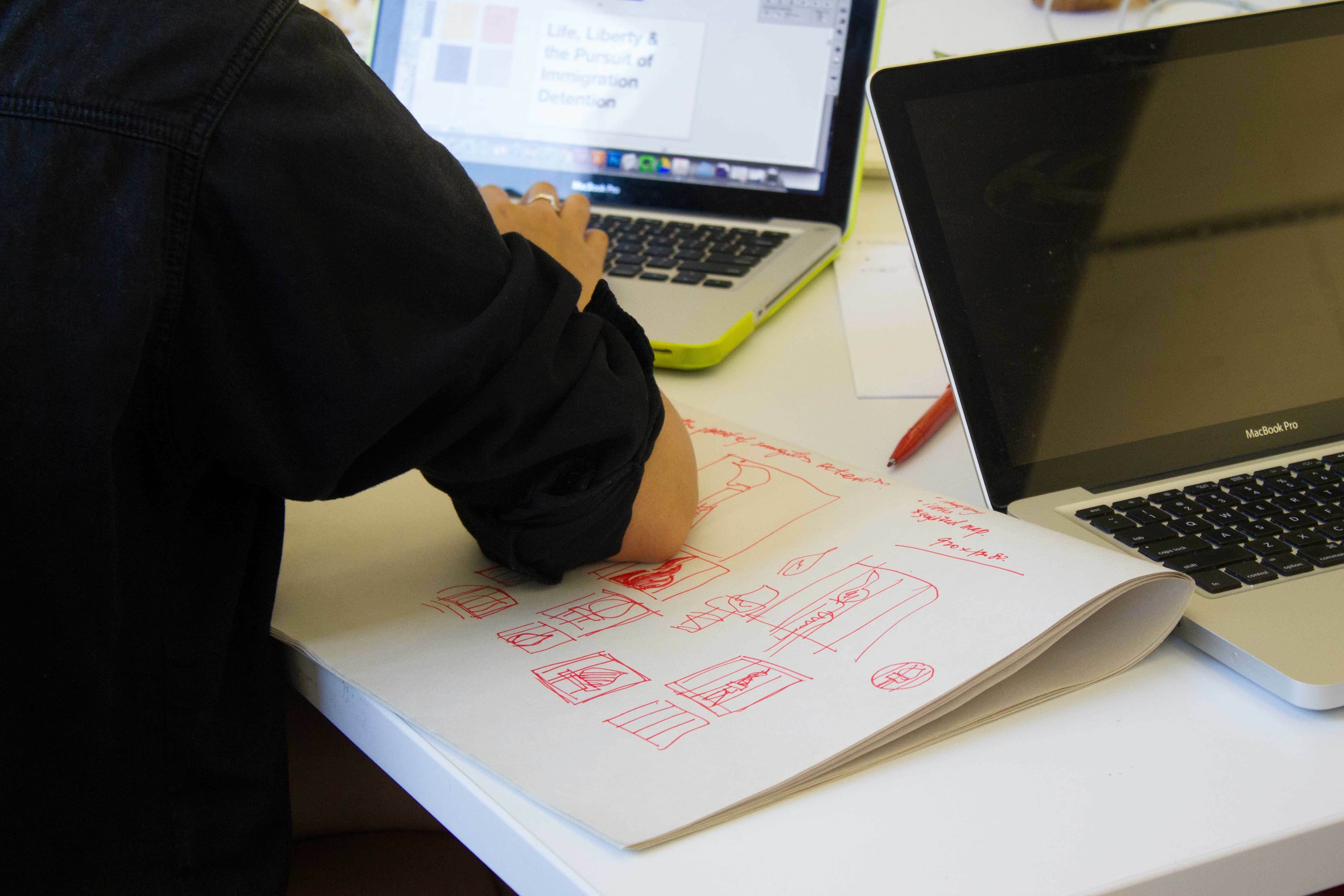 A person is seated at a desk, working between two laptops. Their focus is on a sketchbook with red drawings, presumably planning or designing. One of the laptops displays a document titled "Life, Liberty & the Pursuit of [cut off] Detain [cut off].