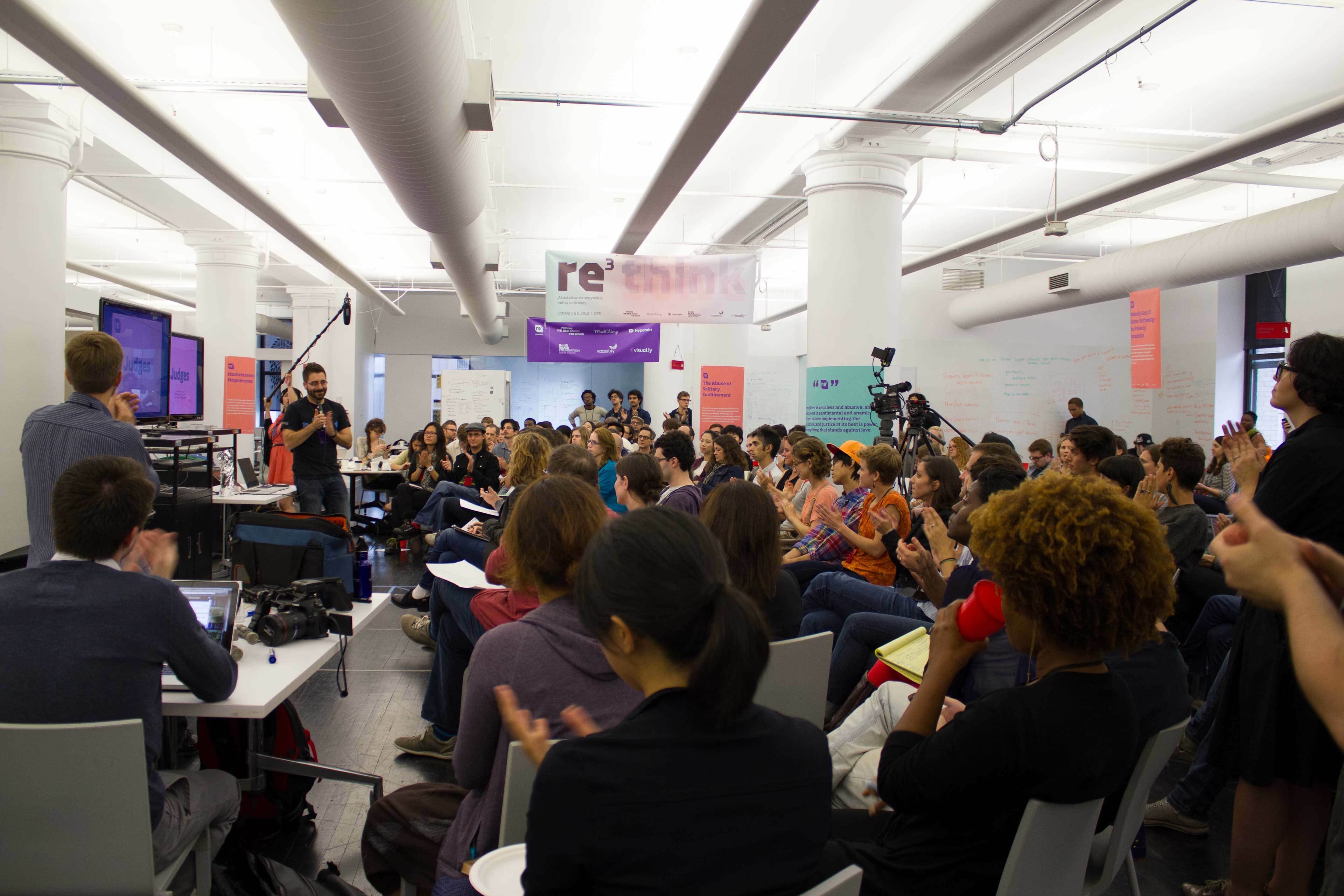 A large group of people is gathered in a modern, open-concept room with white pillars and ceiling ducts. Various individuals are engaged in conversation, taking photos, and listening attentively. A presentation screen is visible in the background, with "re:think" written above.
