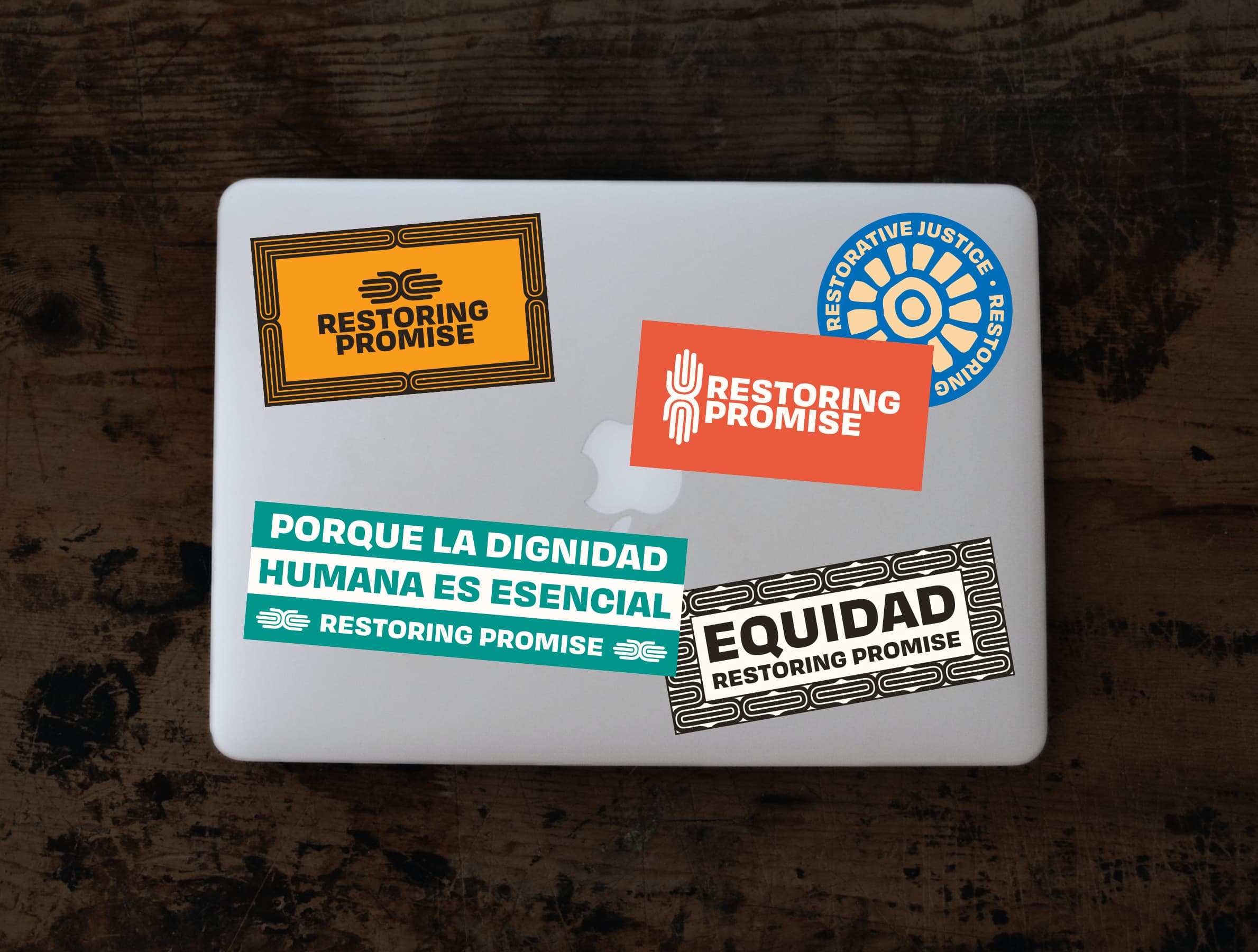 A laptop covered with stickers promoting "Restoring Promise" and restorative justice. The stickers include messages such as "Restoring Promise", "Porque la dignidad humana es esencial", and "Equidad". The laptop sits on a dark wooden surface.