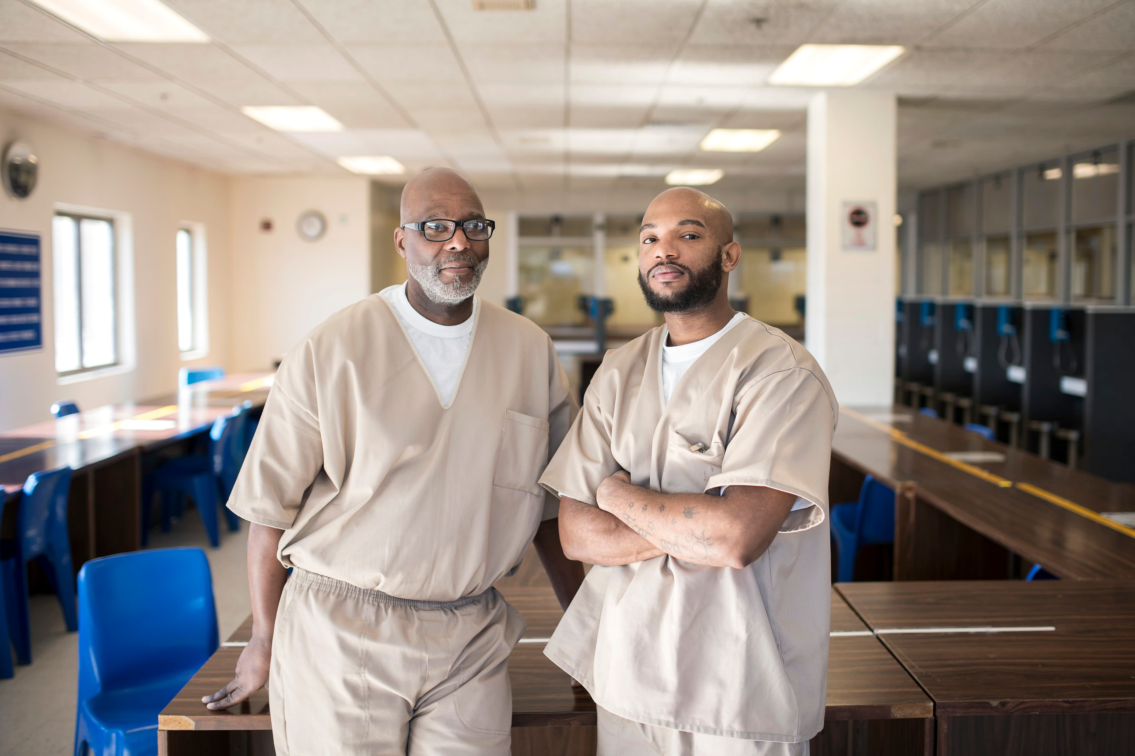 Two men in beige prison uniforms stand side by side in a well-lit common area of a facility. The older man on the left has glasses and a gray beard, while the younger man on the right has a shaved head and is crossing his arms. Blue chairs and tables are seen in the background.