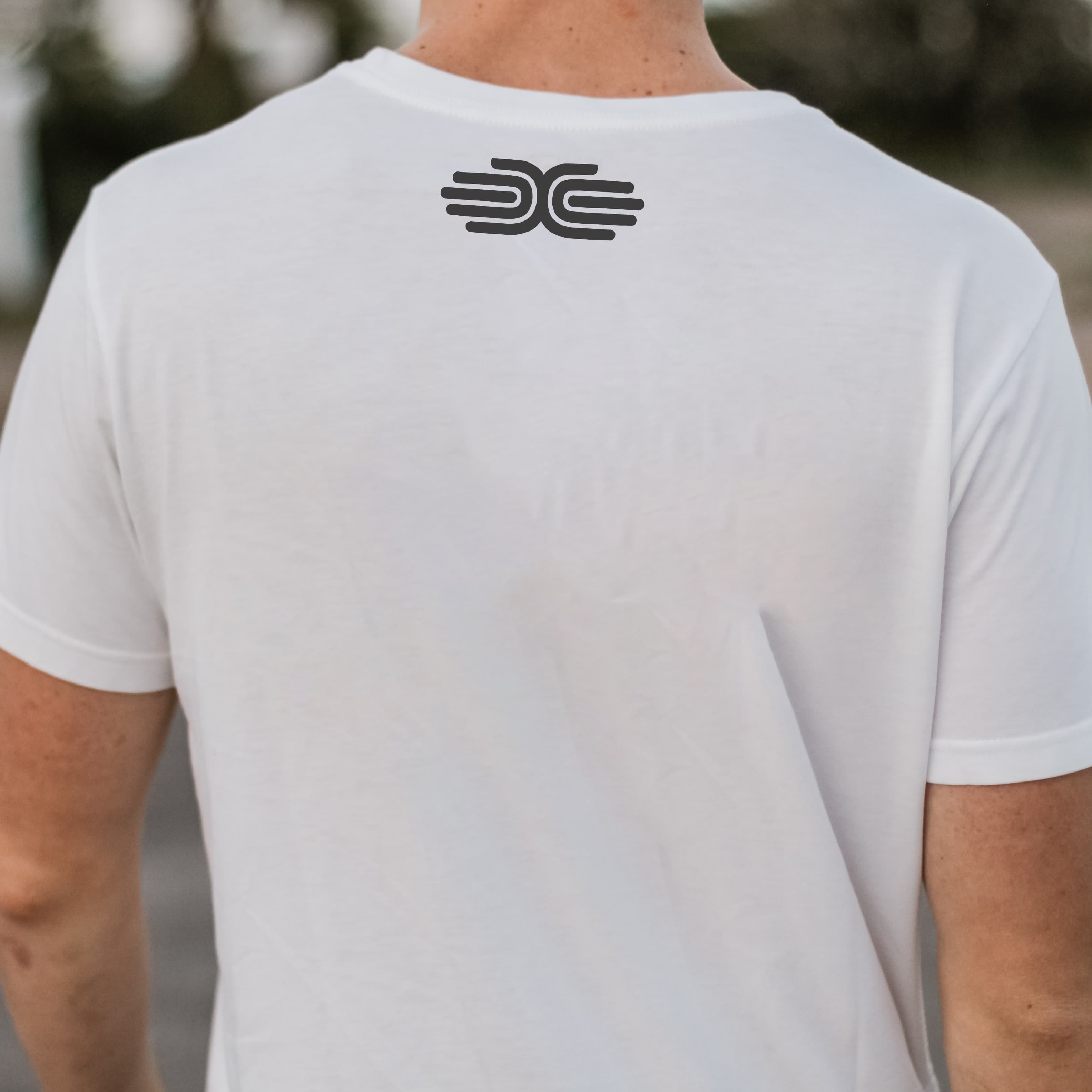 A person wearing a plain white t-shirt is standing with their back to the camera. The t-shirt has a simple black logo on the upper back, consisting of two mirrored, stylized "C" shapes facing each other. The background is blurred and indistinct.