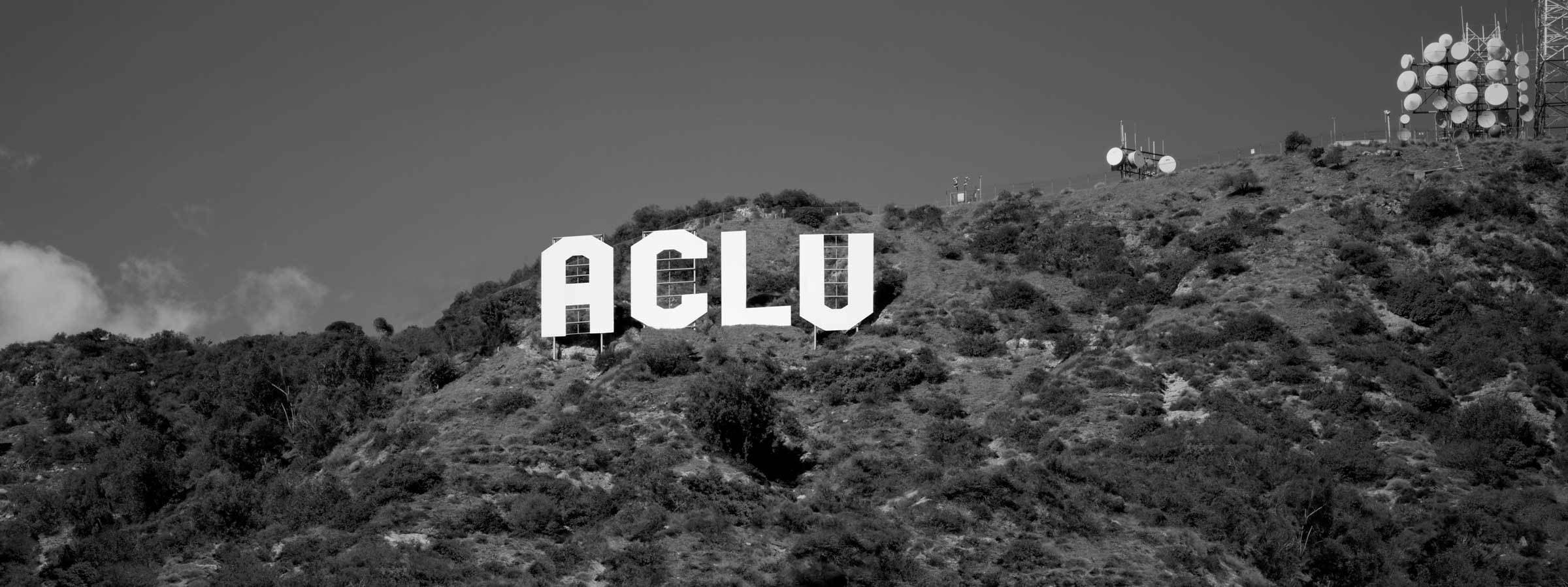 A black and white image shows a hillside with large, white letters spelling "ACLU," reminiscent of the iconic Hollywood sign. The hillside is covered in shrubs and vegetation, with several telecommunications towers visible on the right.