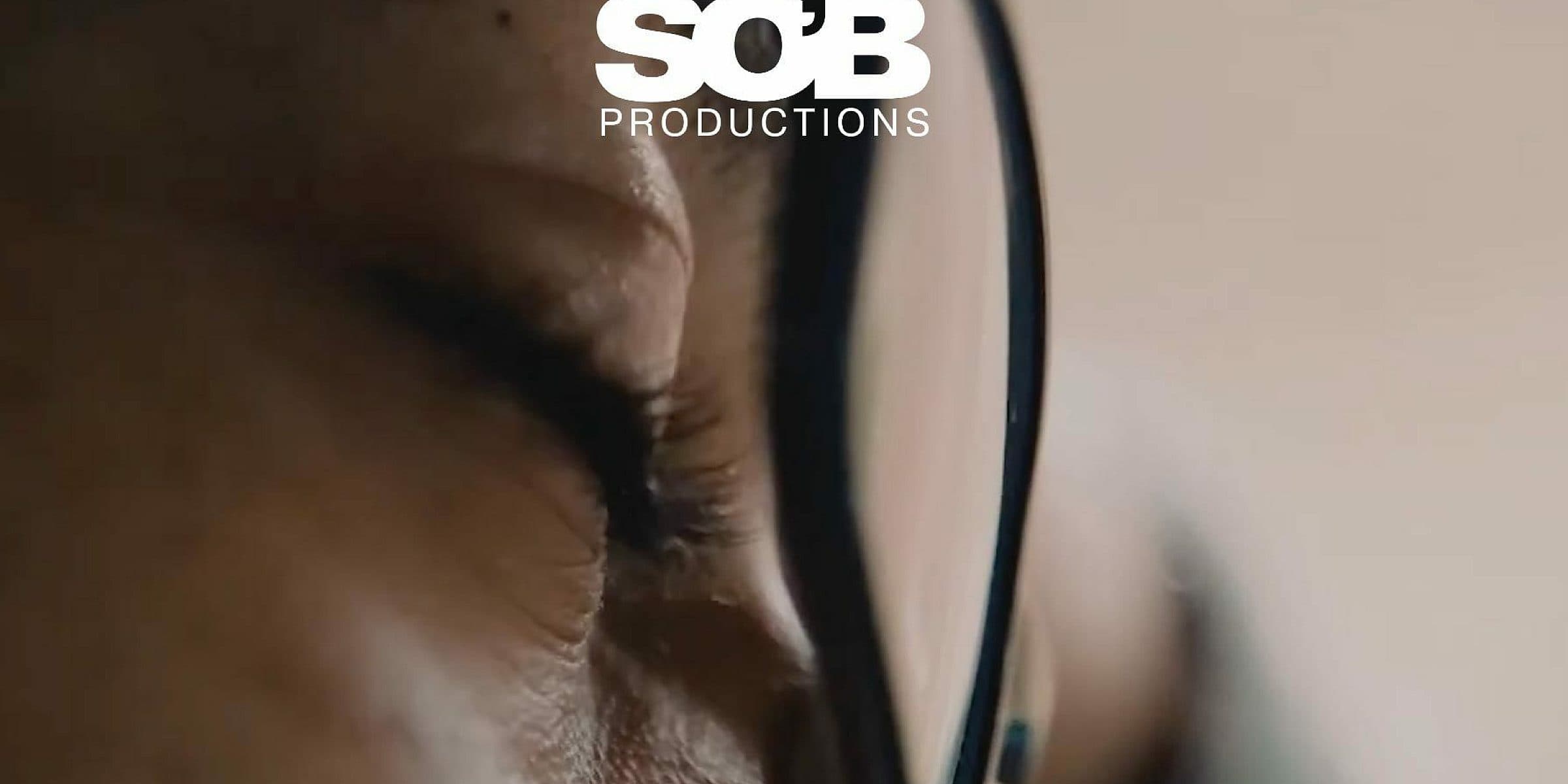 A close-up of a person's face with closed eyes, wearing black-framed glasses. The image has a soft focus, and "SOB Productions" is prominently displayed in white text at the top.