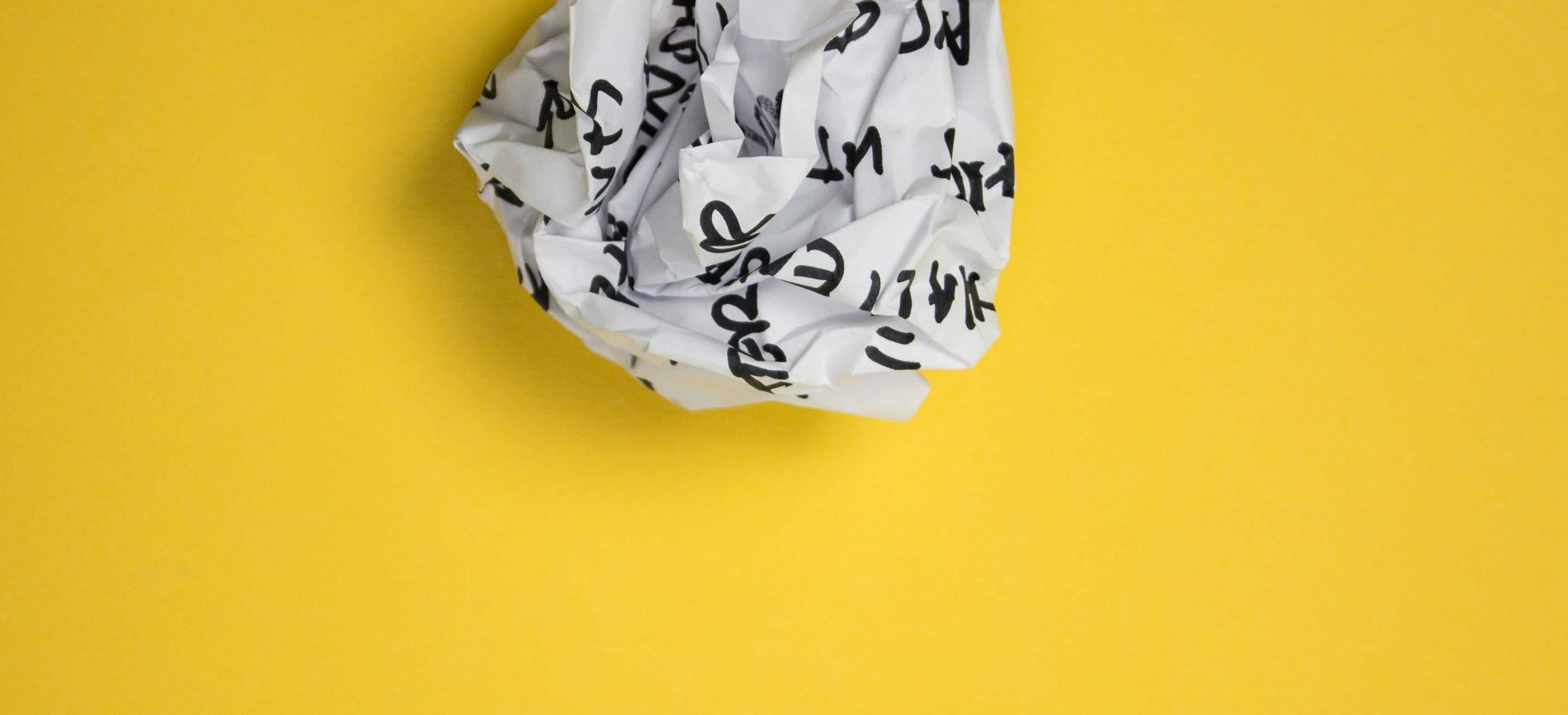 A crumpled piece of white paper with black text on it lies against a bright yellow background. The paper appears to have handwritten or printed characters, but they are not legible due to the crumpling.