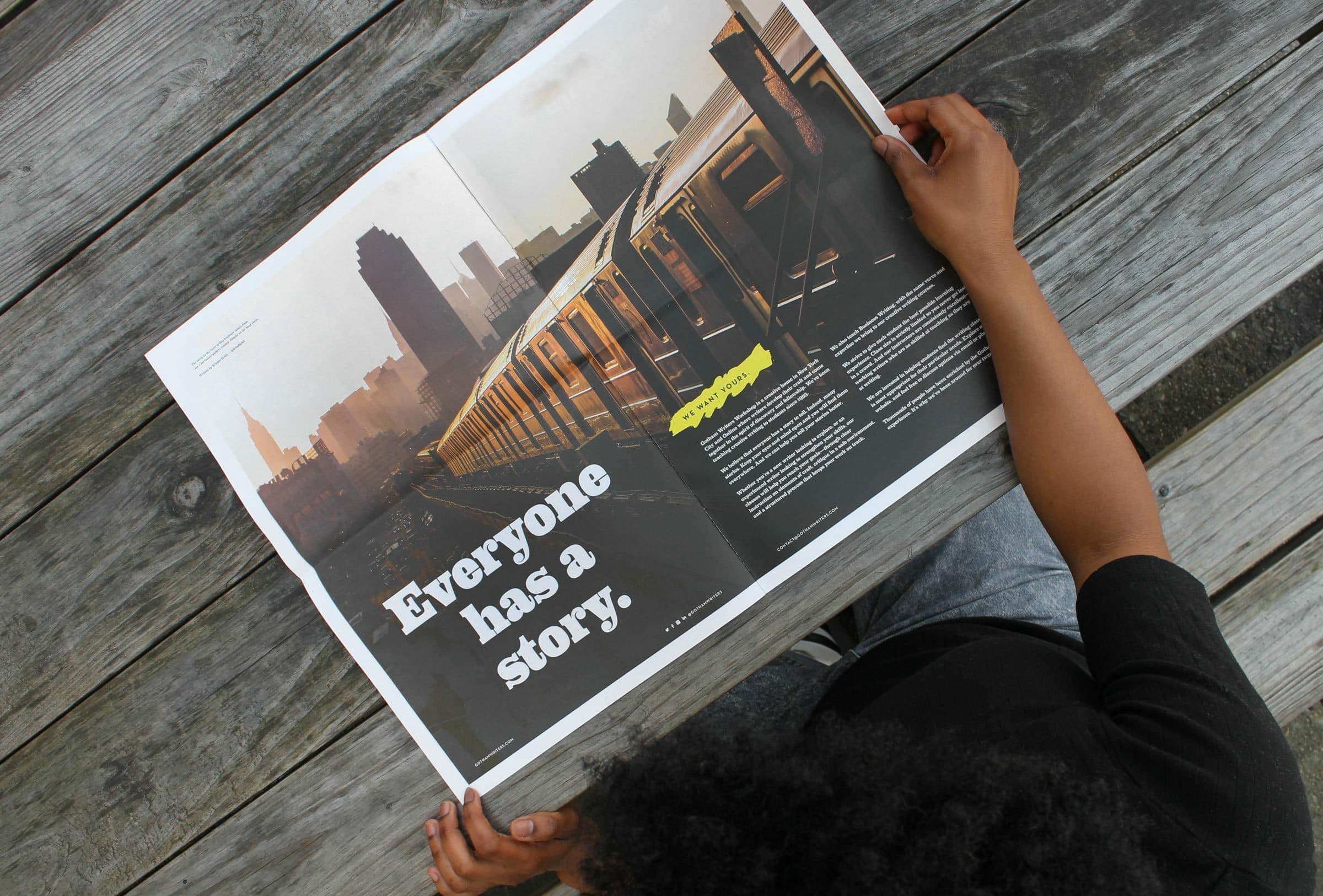 A person with curly hair sits on a wooden surface, holding an open magazine. The left page features a photo of a train and city skyline with the text "Everyone has a story." The right page contains additional text and a small image.