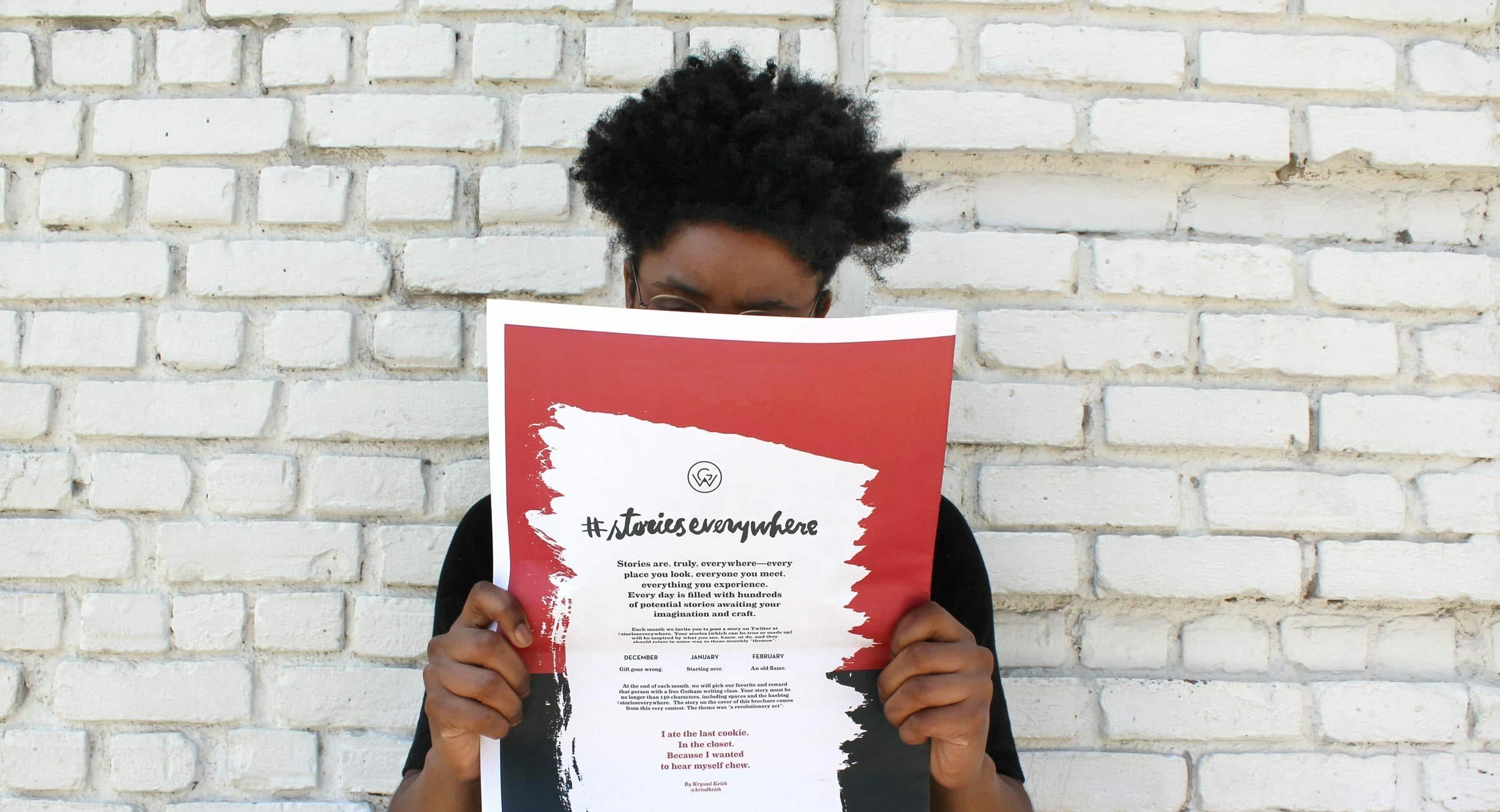 A person with short, curly hair is holding up a red and white paper titled "#storieseverywhere" in front of their face. The person stands against a white brick wall, partially hiding behind the paper which contains text in black and white.