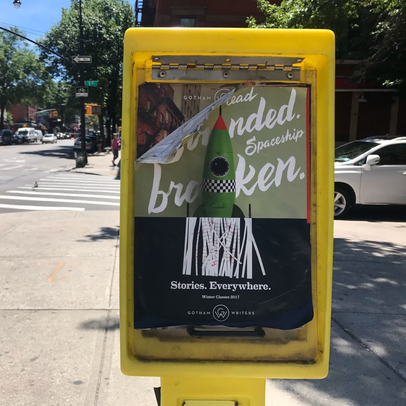 A yellow newspaper box on a city sidewalk features a poster for Gotham Writers with a rocket illustration. The partially peeled poster reveals the words "Spaceship broken. Stories. Everywhere." Trees, buildings, and a crosswalk are visible in the background.