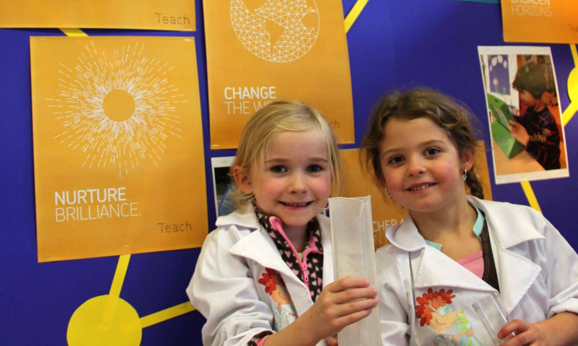 Two young girls wear scientist lab coats while smiling and holding a paper tube in a classroom. Behind them, colorful posters display phrases like "Nurture Brilliance" and "Change the World." The setting appears vibrant and educational with a focus on learning.
