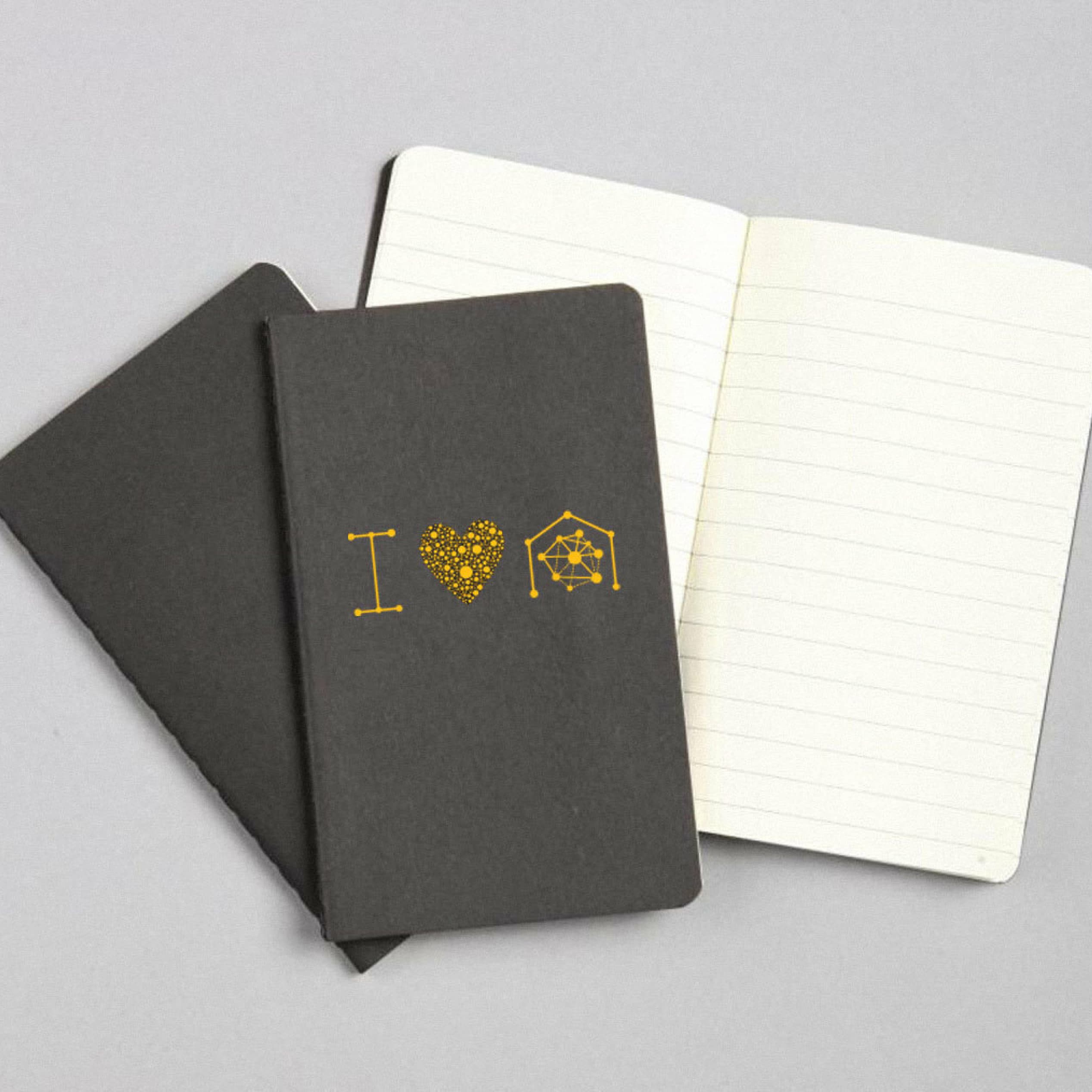 Three notebooks lie on a gray background. Two notebooks are closed, with a black cover featuring a yellow design that reads "I [heart] science" with a heart symbol and an atom. The third notebook is open, showing blank lined pages.
