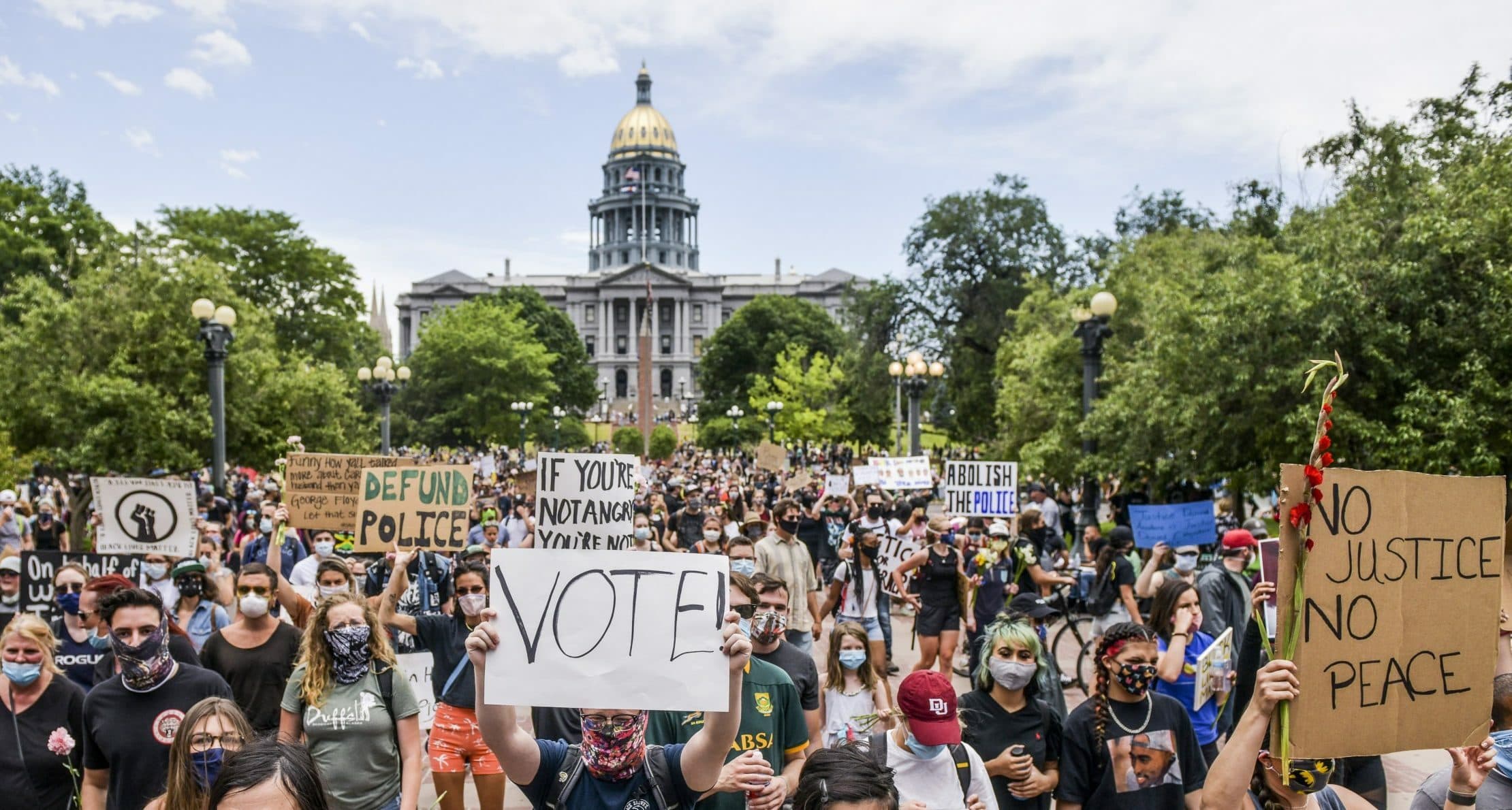 A large crowd of protesters with raised signs gather in front of a government building. The signs include messages such as "Vote!", "Defund Police", and "No Justice No Peace". Many participants are wearing masks, and the sky is partly cloudy.