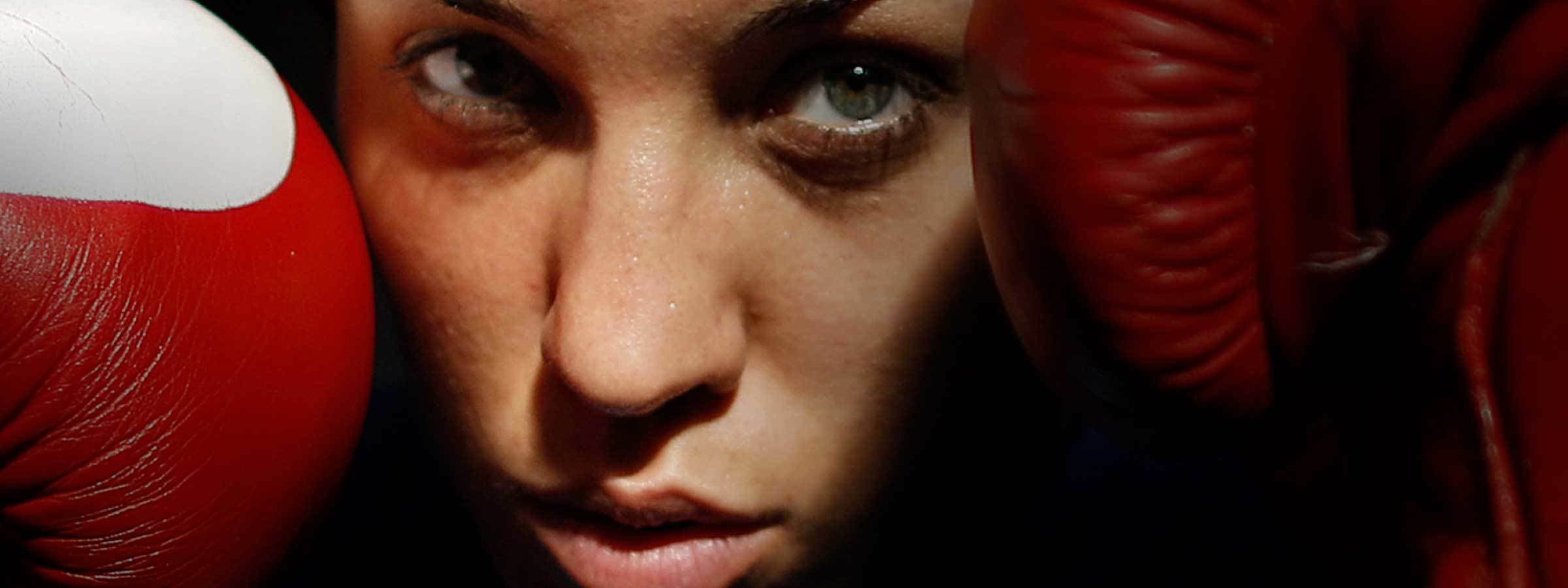 Close-up of a person with intense eyes, framed by two red boxing gloves near their face. The lighting casts a dramatic shadow, highlighting their determined expression.