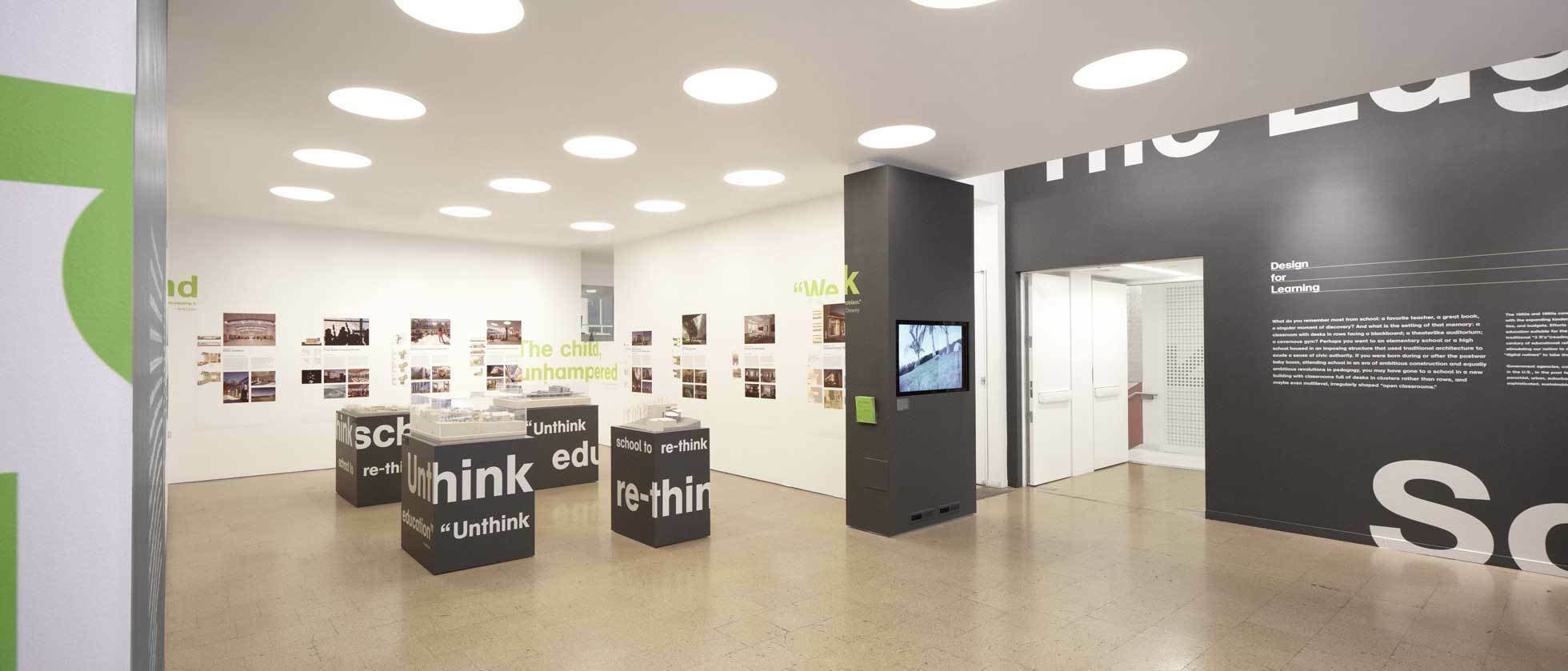 A modern exhibition space with white walls and light wood floors. Black columns display text and images. The walls feature text and photo displays, and there are text-covered rectangular display stands in the center. Circular ceiling lights illuminate the room.