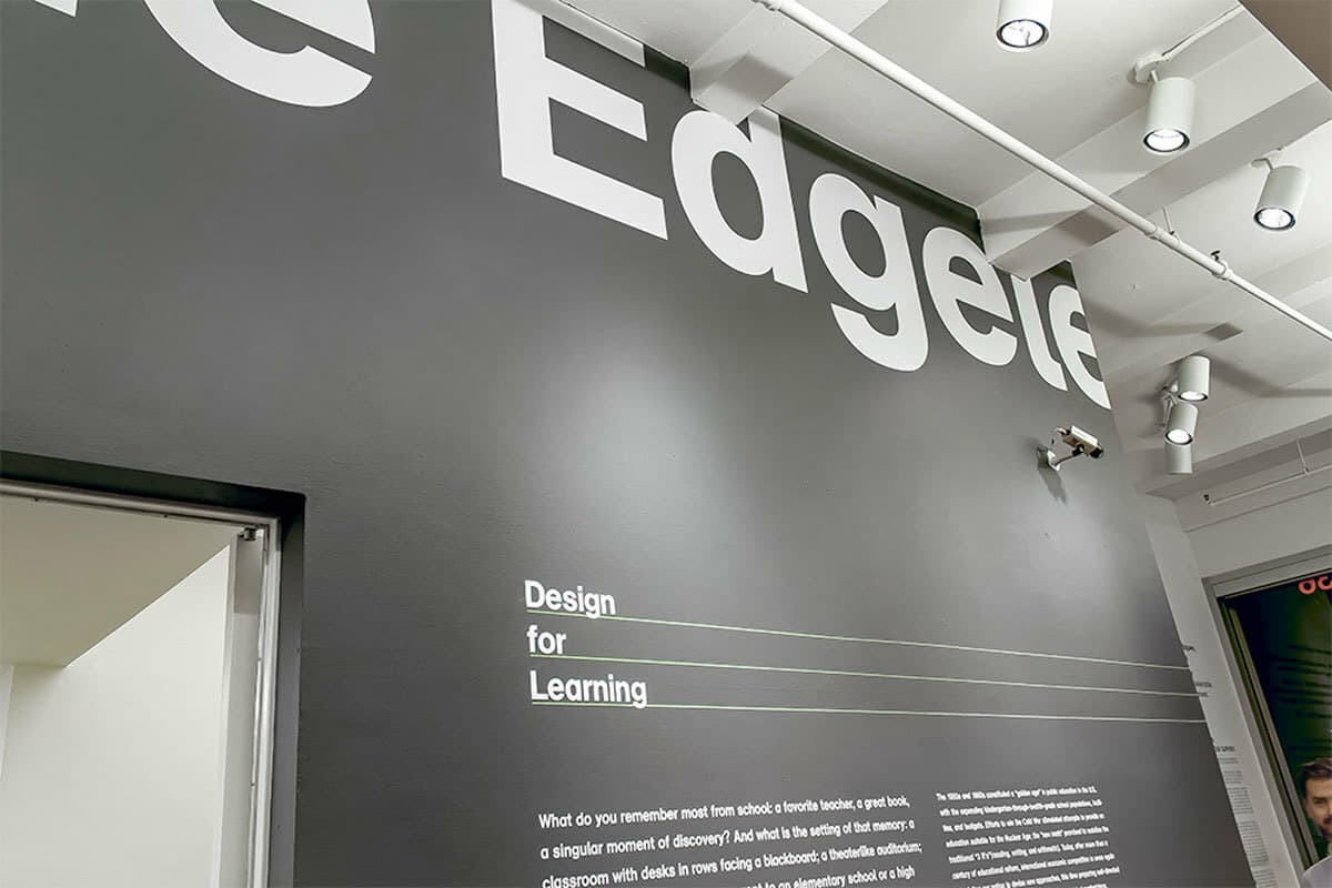 A modern interior space with a black wall featuring large, white text that starts with "Edge". Smaller white text below reads "Design for Learning". The ceiling has exposed beams and multiple spotlights. A security camera is mounted on the wall.