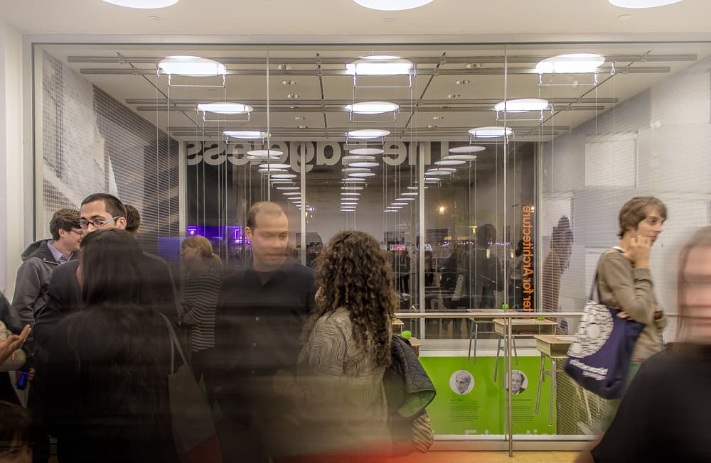 A busy indoor scene with people walking and chatting in a glass-fronted area. A man in a black shirt and a woman with curly hair are near the middle. Some people are blurred due to movement. In the background, a modern-lit area is visible with text on the walls.