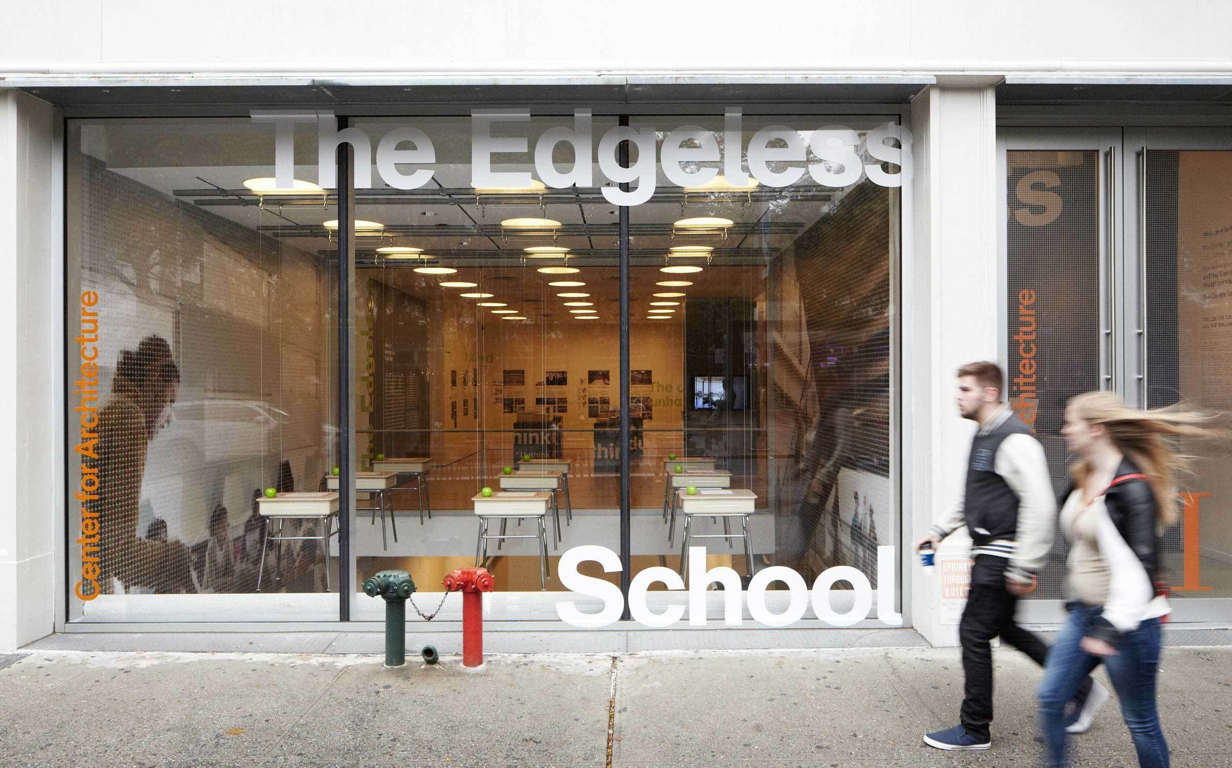 Two people walk past the glass windows of a storefront labeled "The Edgeless School" and "Center for Architecture." Inside, tables and chairs are visible, with floor-to-ceiling curtains and ceiling lights. The sidewalk in front has two decorative fire hydrants.