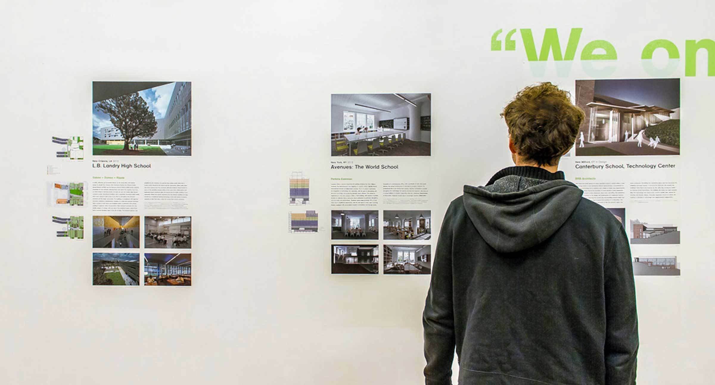 A person in a dark hoodie stands in front of a gallery wall, looking at three architectural display boards. Each board has images and text about different school buildings. Part of a green wall text reading "We on" is visible.