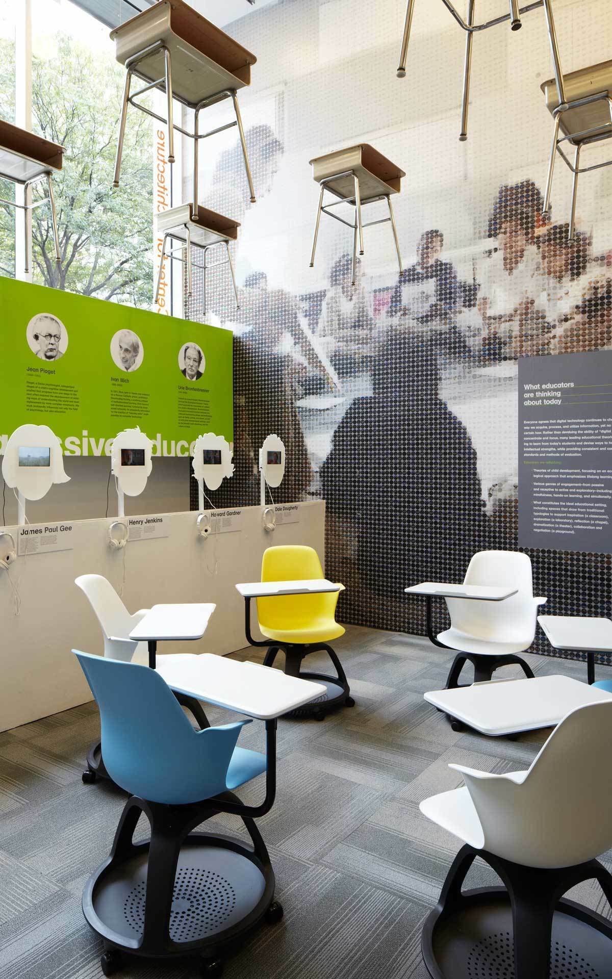 A modern classroom setup with various colorful swivel chairs on wheels, each equipped with a small attached desk. The background features a large mural of students and a green wall with historical figures' images. Some chairs are mounted high on a wall display.