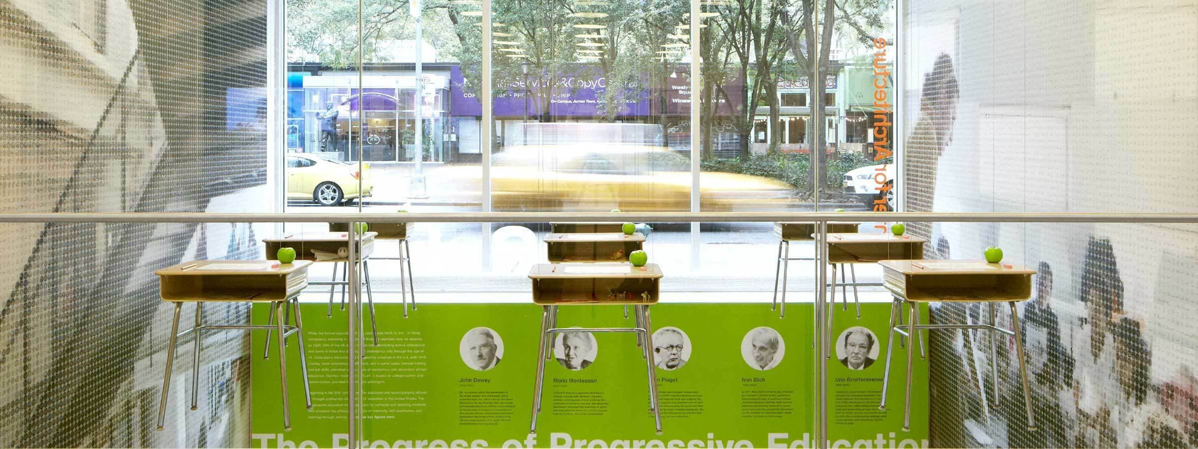 A modern cafe with tall windows showcasing a busy street outside. The interior features high tables with green apples on them and stools arranged neatly. A green panel on the wall displays text and photographs of notable figures related to progressive education.