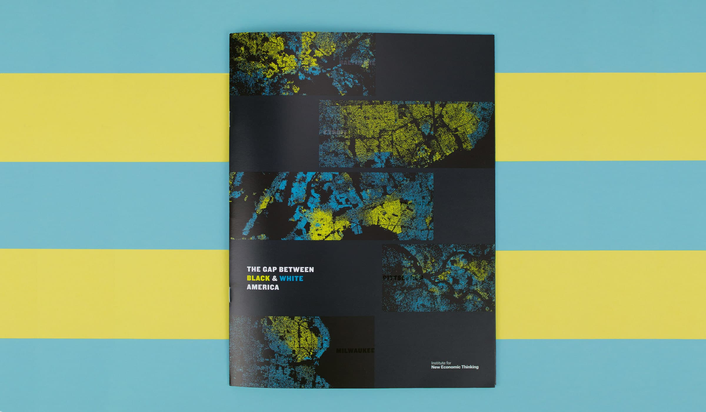 A booklet titled "The Gap Between Black & White America" is placed on a blue and yellow striped background. The cover features abstract, colorful patterns with sections in yellow, green, and blue. The text "MILWAUKEE" and "Inspired by New Economic Thinking" are also visible.
