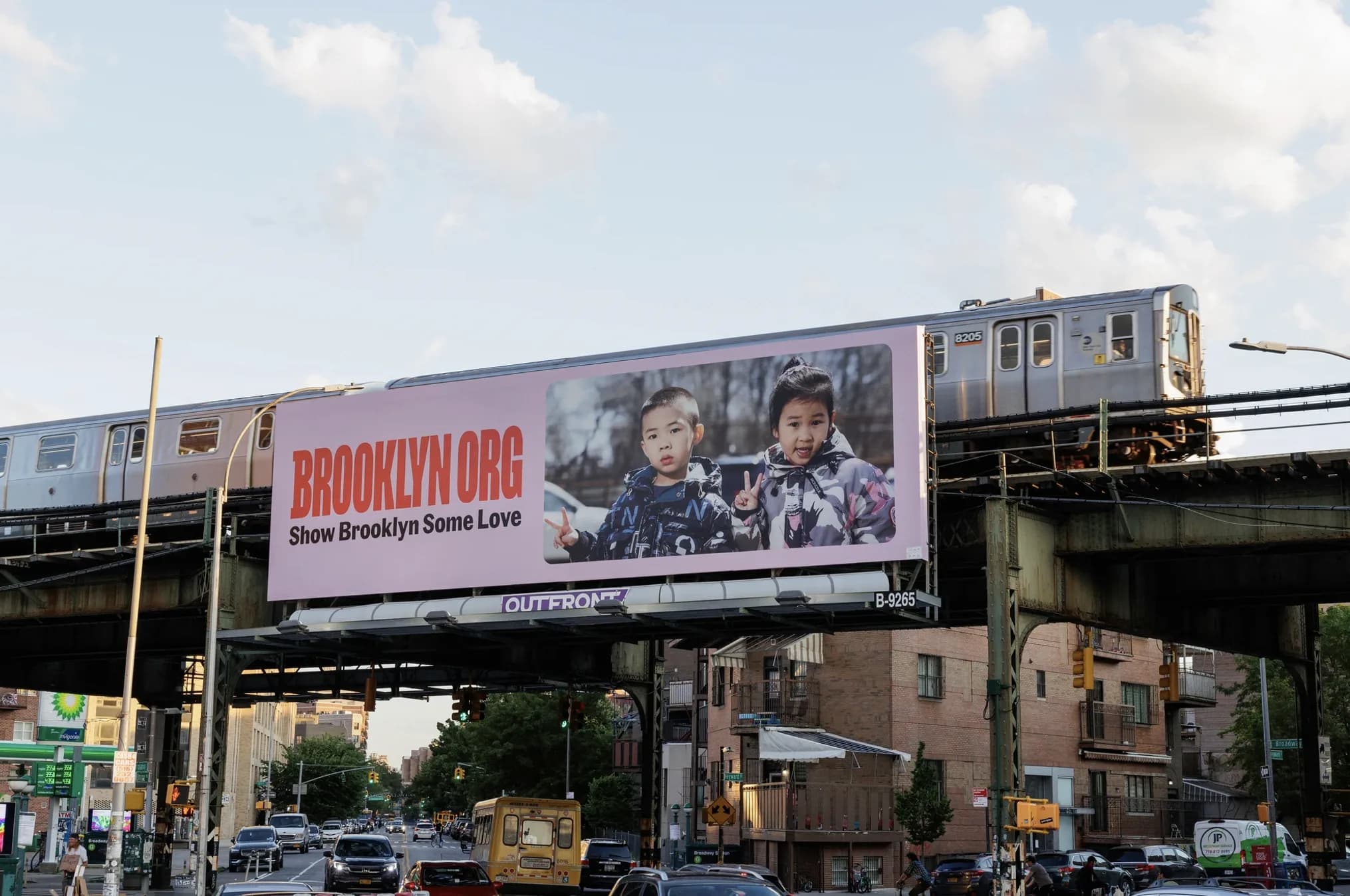 A billboard on an elevated train platform shows two children posing and smiling, with text reading "BROOKLYN.ORG Show Brooklyn Some Love." A subway train passes above, and traffic moves below on a city street.