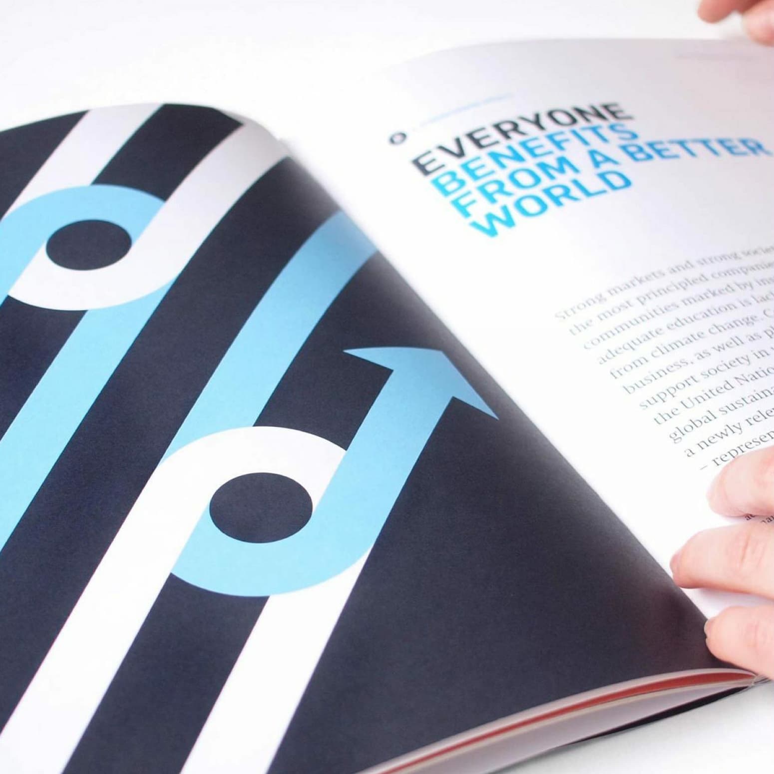 Close-up of an open booklet. The left page features a graphic with curved blue arrows on a black background with white diagonal stripes. The right page contains text starting with the headline "Everyone Benefits from a Better World." Two hands hold the booklet open.