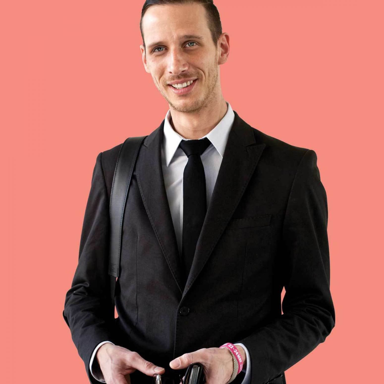 A man wearing a black suit and tie stands against a solid coral background. He is smiling and holding a wallet in his hands. He has a black shoulder bag strap visible over his suit and a pink wristband on his left arm.