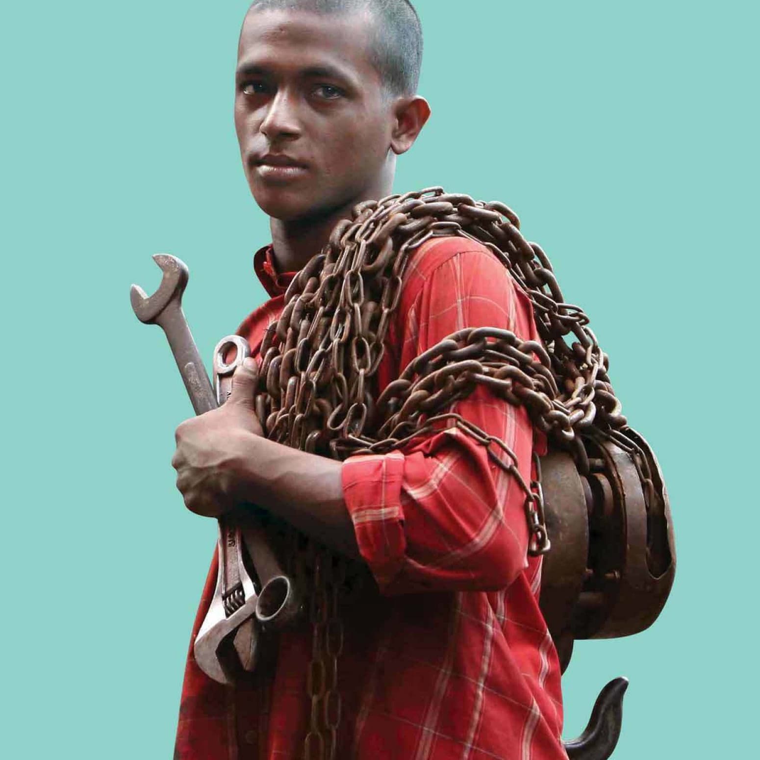 A man in a red plaid shirt is looking at the camera while holding large wrenches and chains over his shoulder against a plain teal background.
