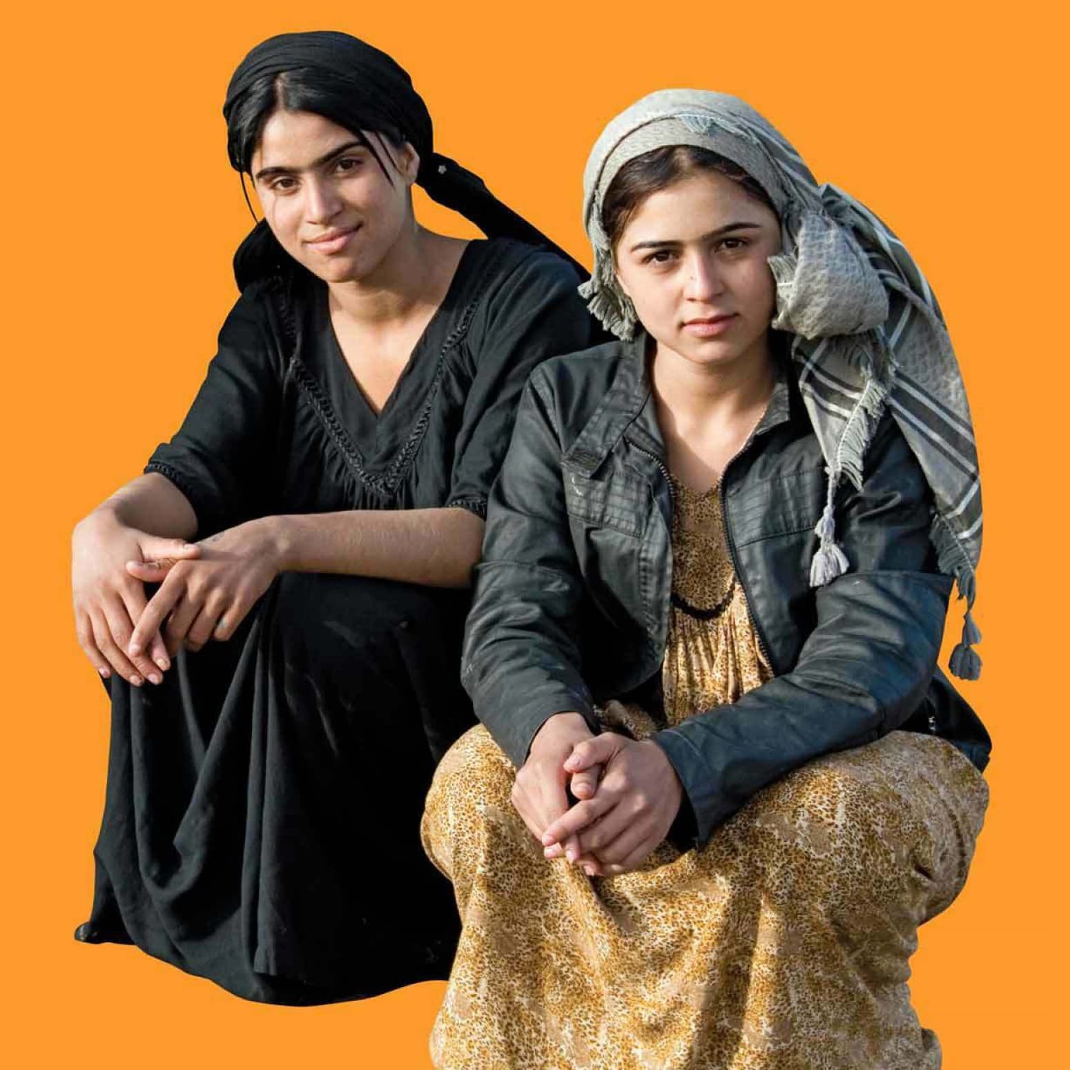 Two women dressed in traditional clothing sit side by side against an orange background. One wears a black dress with a black headscarf, while the other wears a brown patterned dress with a black jacket and a checkered headscarf. Both look directly at the camera.