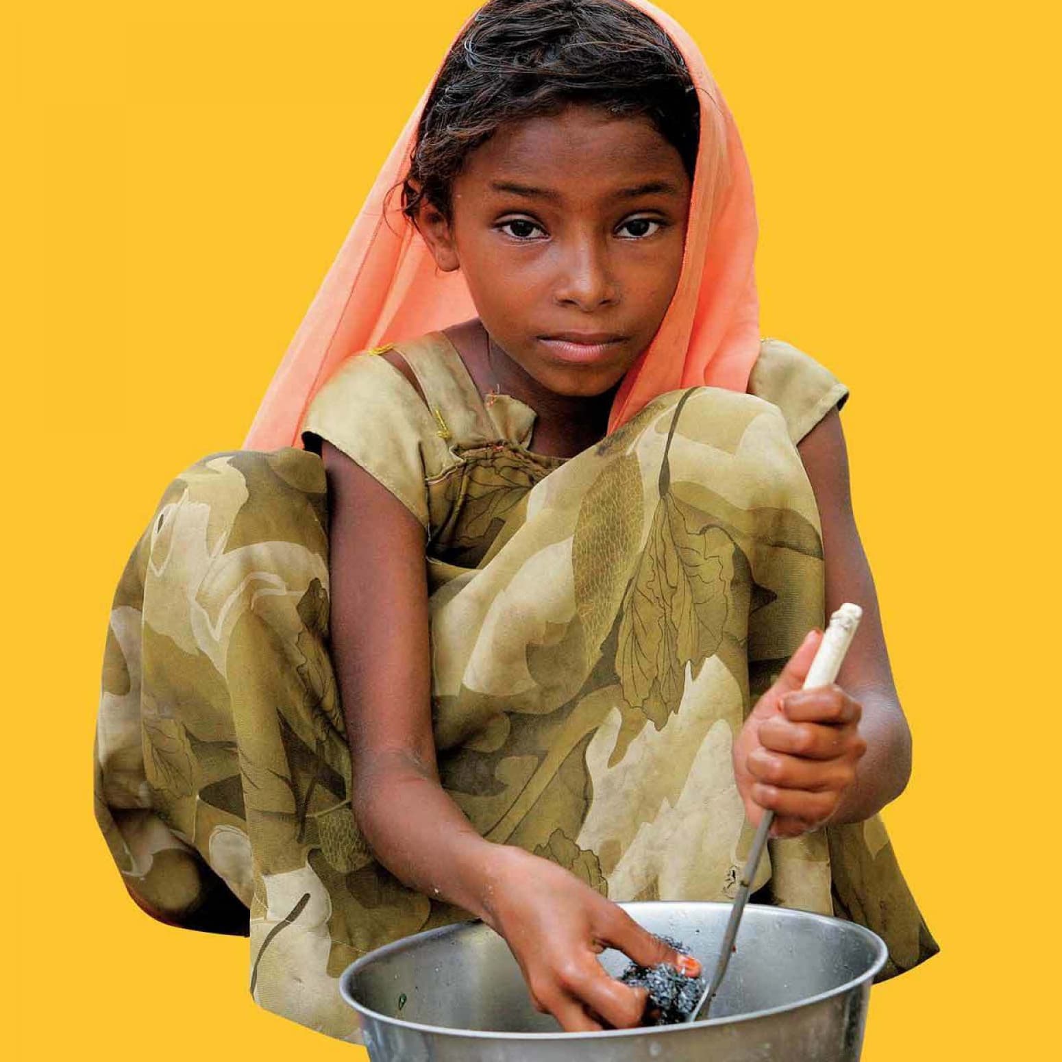 A young girl with brown skin and dark hair, wearing a camouflage-patterned dress and an orange headscarf, is seated and looking at the camera while washing a metal bowl against a bright yellow background.