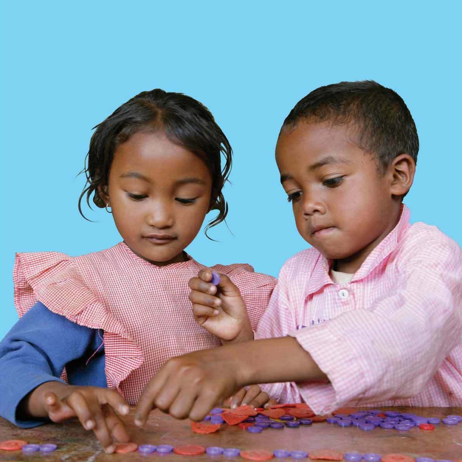 Two children sitting at a table, concentrating on arranging colorful circular pieces. They are dressed in pink and blue outfits, set against a plain blue background. The girl has a pink gingham dress with ruffled sleeves, while the boy wears a pink shirt.