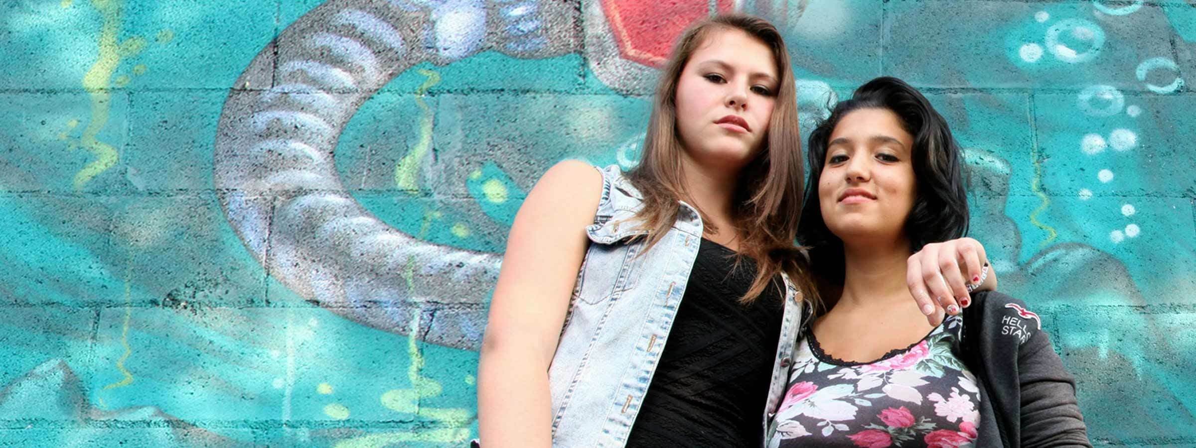 Two young women stand in front of a colorful graffiti wall. The woman on the left wears a denim vest and has long, light brown hair, while the woman on the right has shoulder-length dark hair and wears a floral top. Both look confidently at the camera.