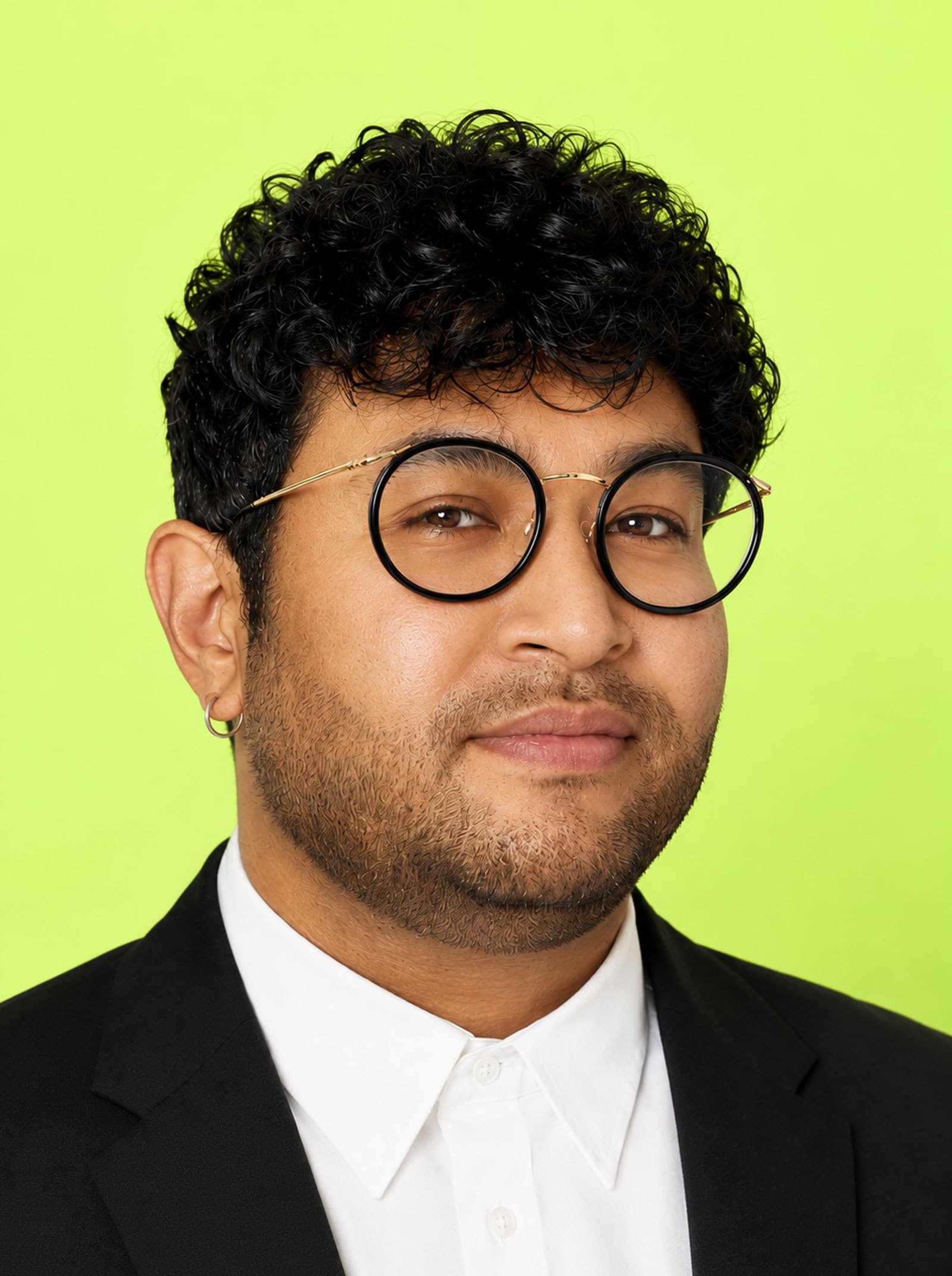 A person with short curly hair, wearing round glasses, a black suit, and a white shirt, poses against a bright green background.