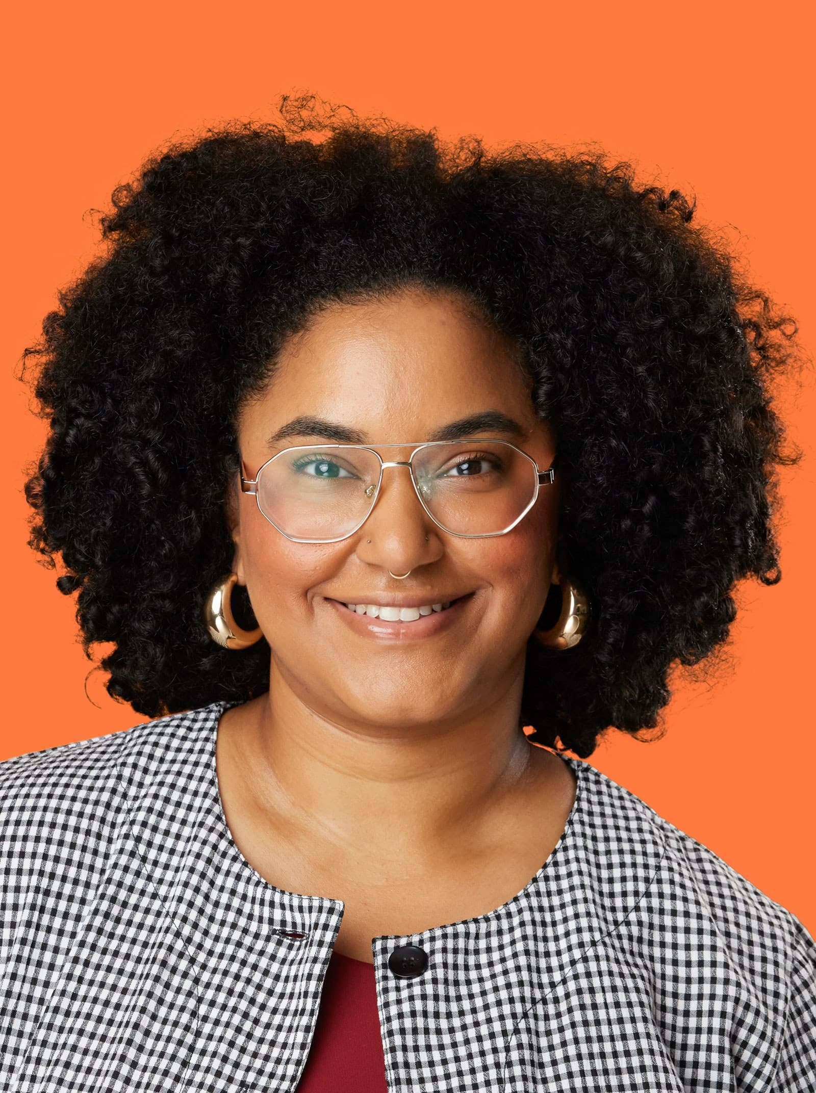 A person with curly black hair, wearing glasses, large gold hoop earrings, and a checked jacket over a burgundy top, smiling in front of an orange background.
