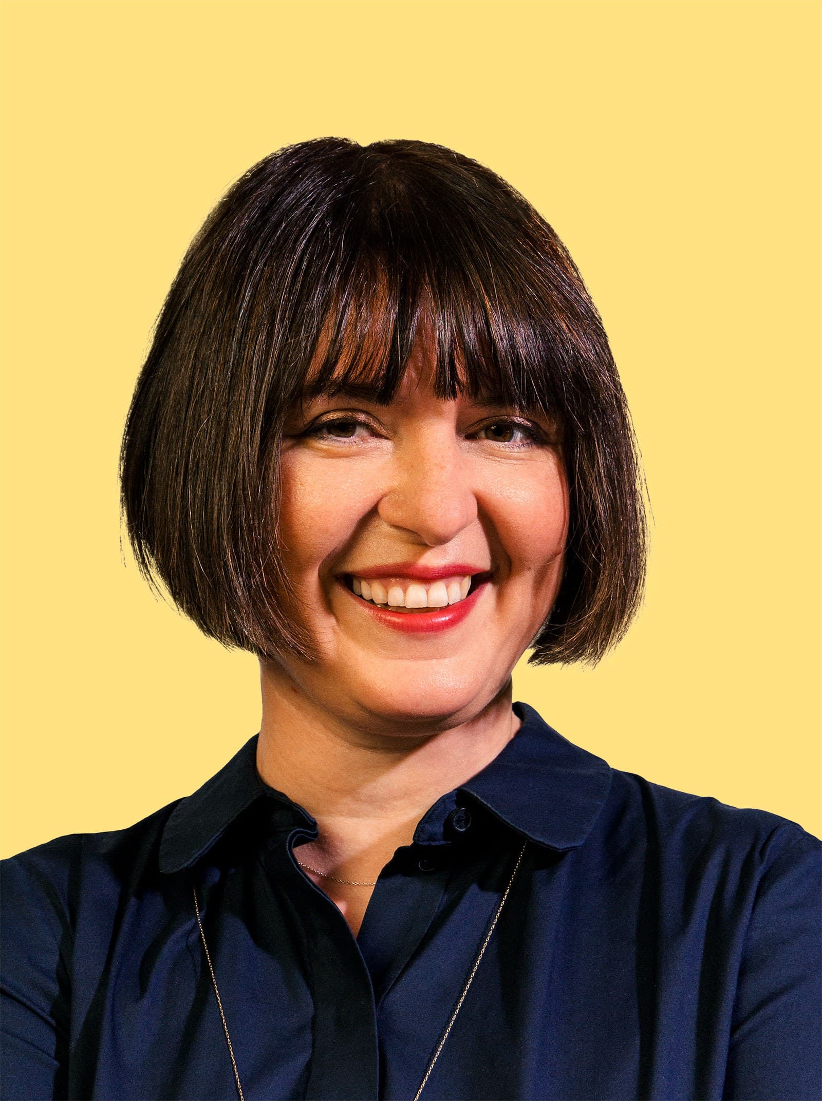A woman with a short dark bob haircut and bangs, wearing a navy blue collared shirt, smiles warmly at the camera against a plain yellow background.