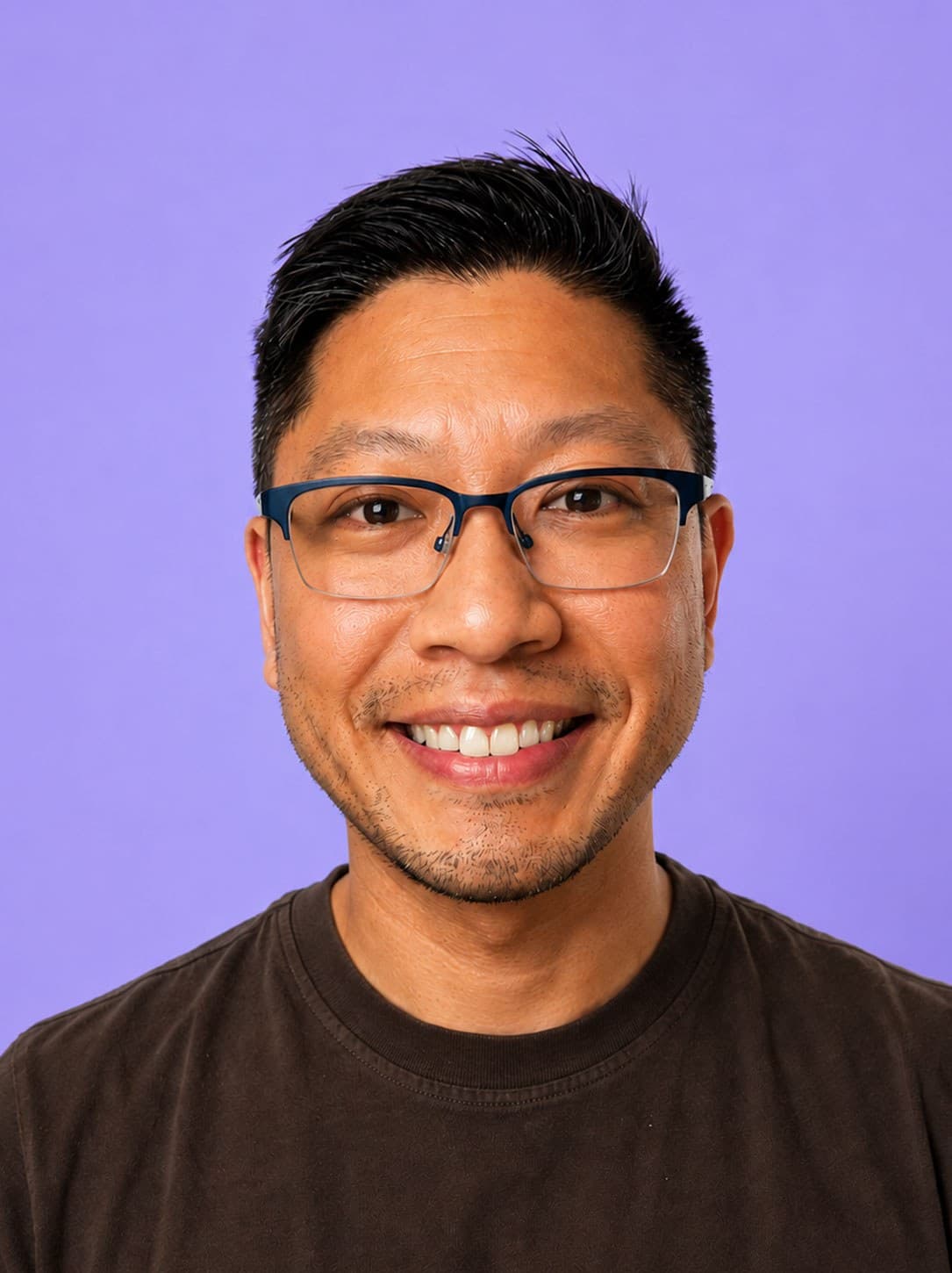 A man wearing glasses and a dark t-shirt smiles at the camera in front of a solid purple background.