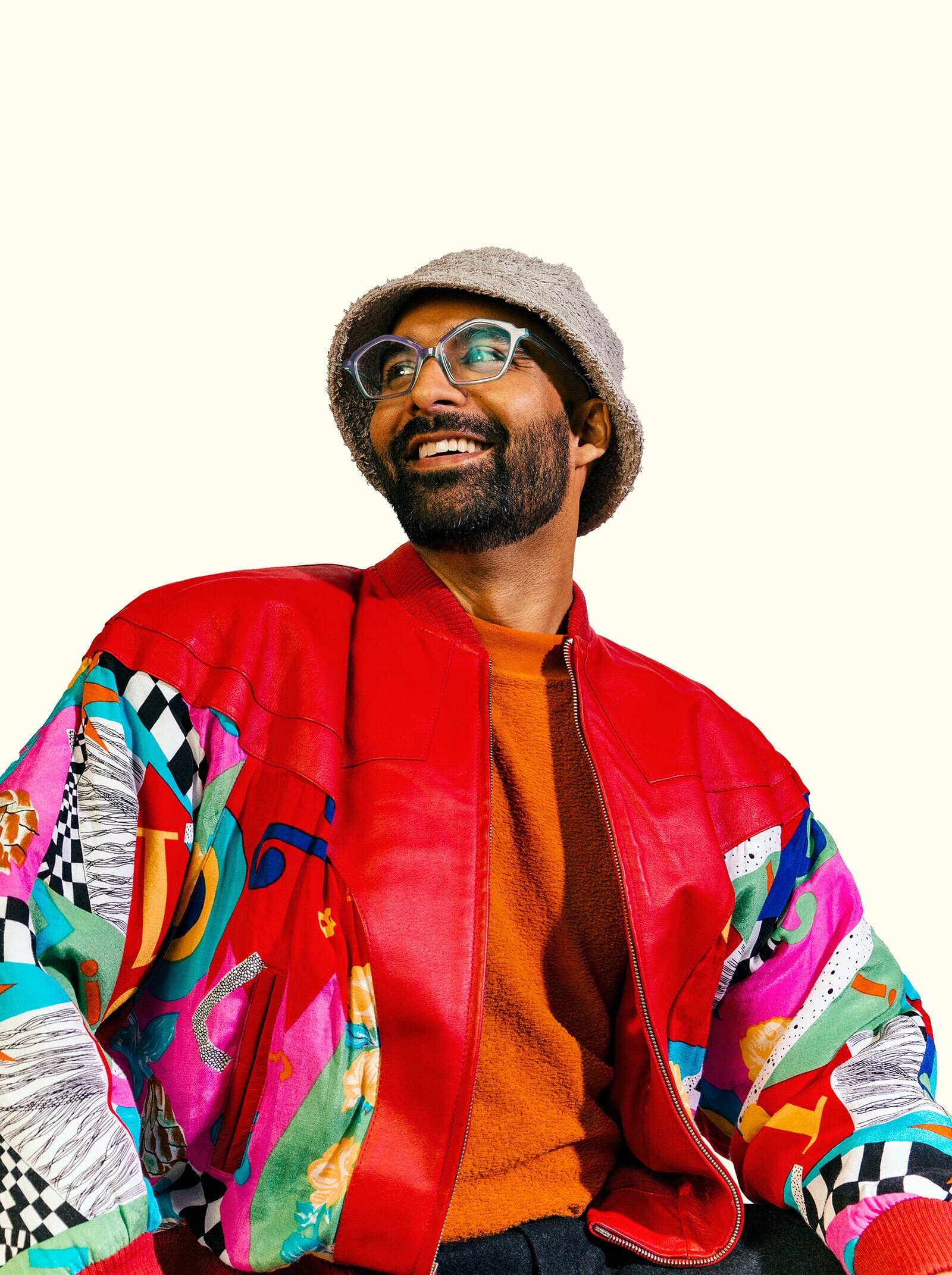 A person with a beard and glasses smiles and looks to the side. They are wearing a colorful, patterned jacket, a textured hat, and an orange top. The background is a plain, light color.