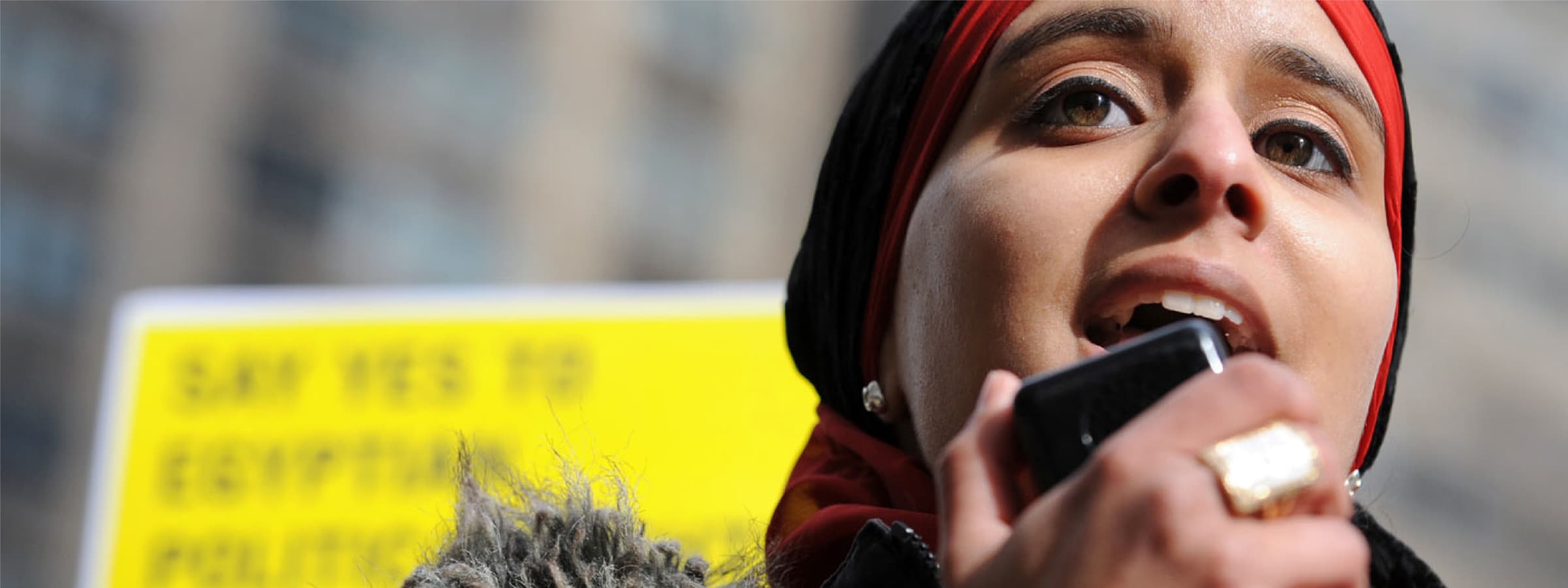 A person wearing a red and black headscarf speaks into a microphone. They are outdoors in front of a blurred background with a yellow sign partially visible, displaying text that starts with "SAY YES TO EGYPTIAN." The individual looks determined and focused.