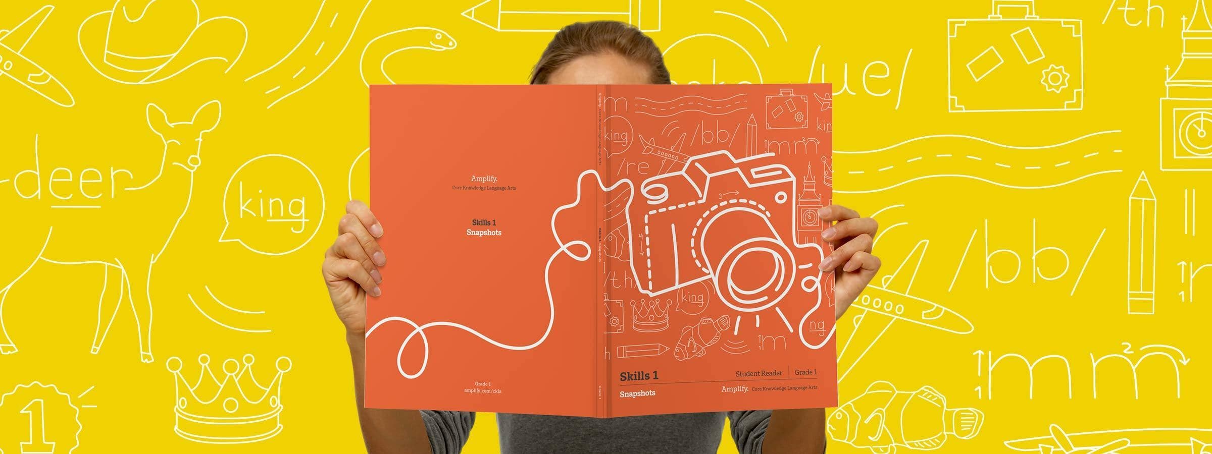 A person holds a red booklet featuring a white outline drawing of a camera and various objects. The background is yellow with white doodles of items such as a suitcase, a cactus, a crown, and the word "king." The person's face is obscured by the booklet.