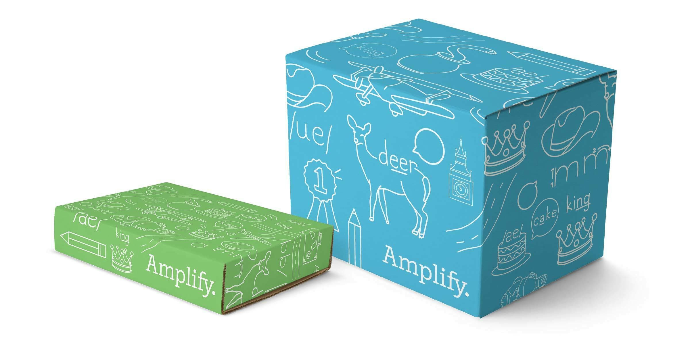 Two cardboard boxes with colorful, playful designs and the word "Amplify" printed on them. The larger box is blue, adorned with white doodles including a deer, cake, and other whimsical items. The smaller box is green, featuring similar doodles.