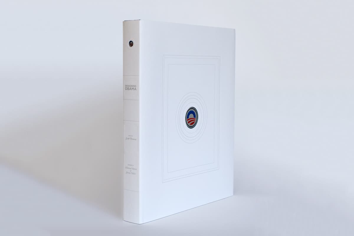 A large white hardcover book stands upright with a minimalist design. The cover features a small emblem in the center, resembling a simplified logo. The spine of the book has text that appears to read "Designing Obama.