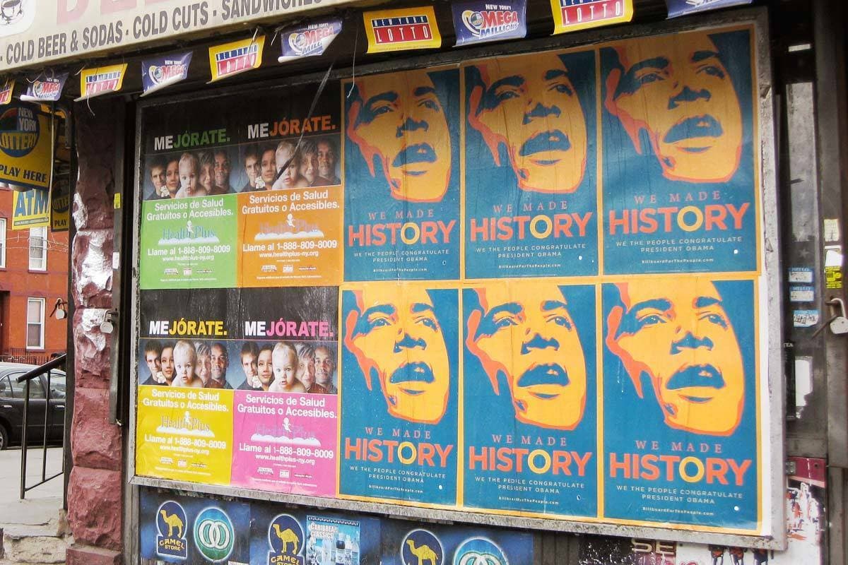 Posters cover a storefront window. Most of them display a stylized image of a person with the text "WE MADE HISTORY" beneath it. Other posters promote health services. The storefront's awning advertises beer, sodas, and sandwiches.