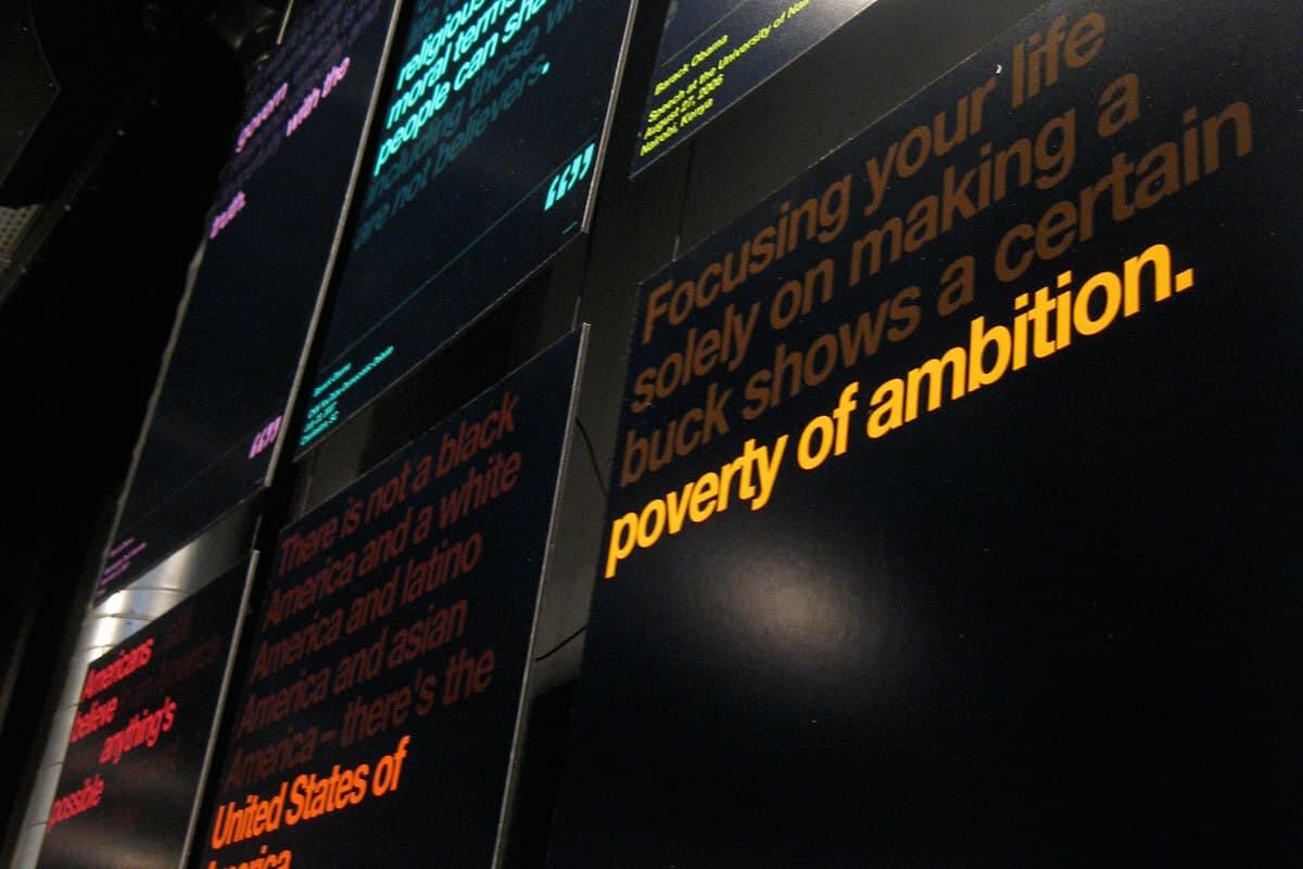 Close-up of multiple digital screens displaying various quotes in different colors. The closest screen shows the quote "Focusing your life solely on making a buck shows a certain poverty of ambition" in orange text on a black background. Other quotes are partially visible.