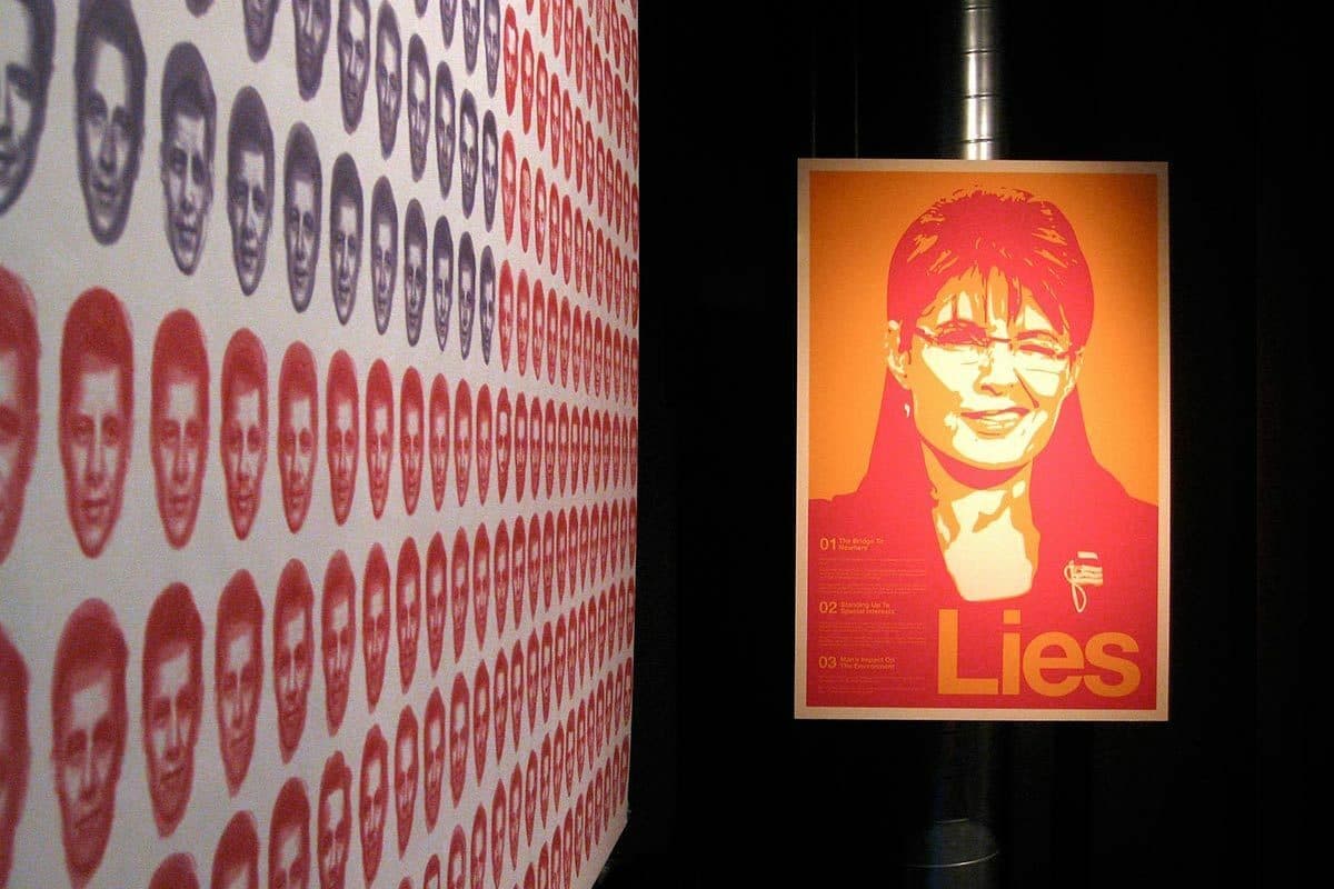 The image features a wall adorned with numerous small, monochromatic visage prints. Adjacent, a large poster displays a stylized, orange-toned portrait of a woman with glasses and the word "Lies" in bold letters beneath her image.