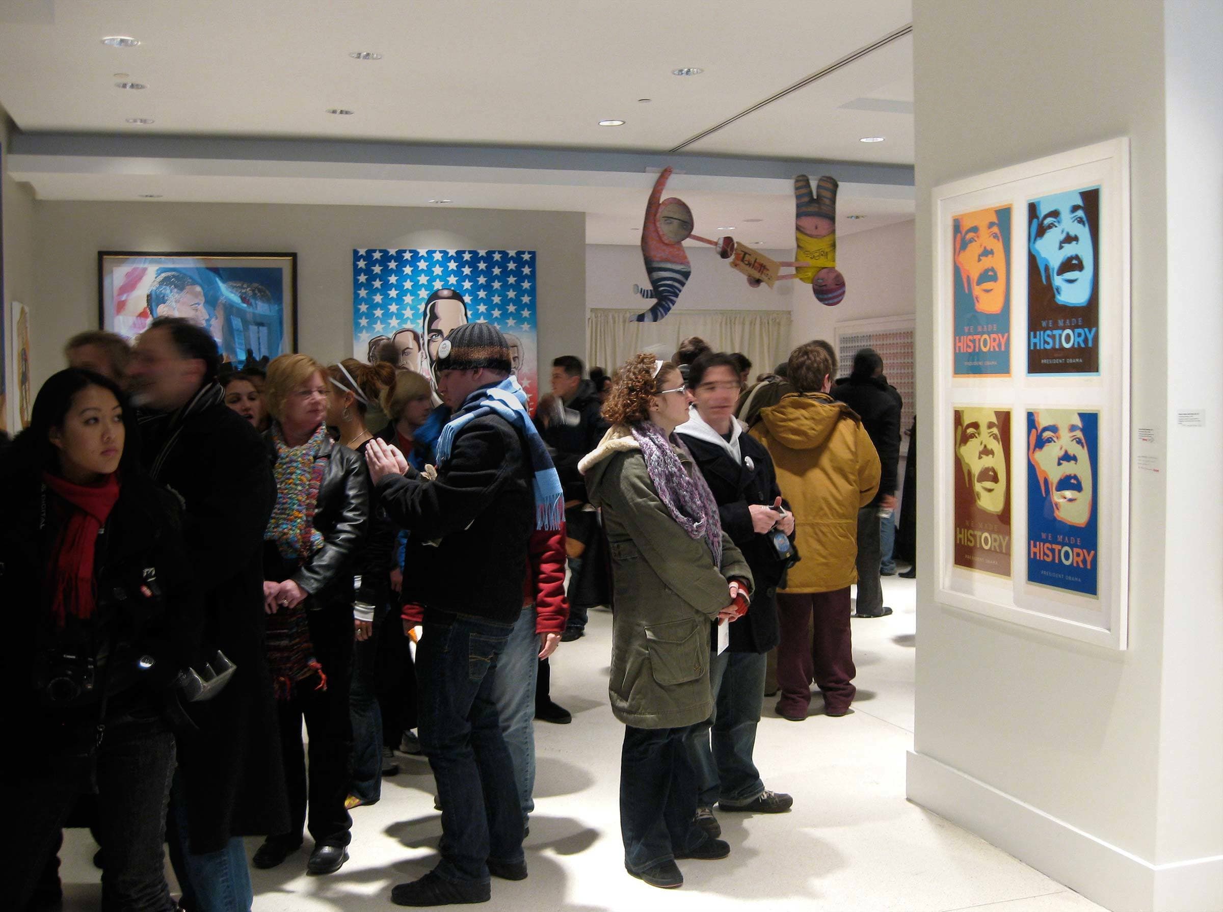 A group of people stands in an art gallery, observing and discussing various artworks. The gallery walls display colorful, framed pieces, including a series of posters with the word "HISTORY" and stylized portraits. The room is brightly lit and modern.