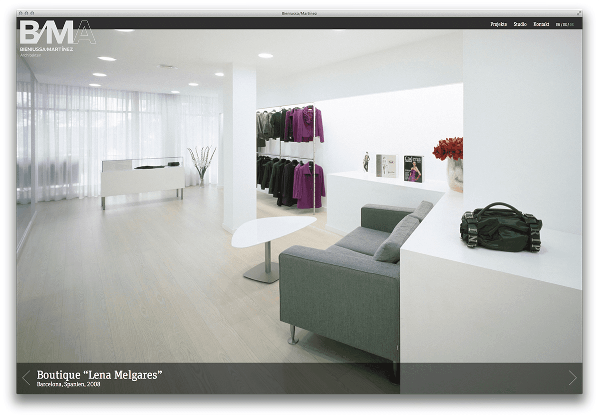 Modern boutique interior with light wood flooring, white walls, and minimalist decor. Features a gray sofa, white tables, and shelves displaying colorful clothing. A sleek white reception desk is in the background, and large windows with sheer curtains allow natural light.