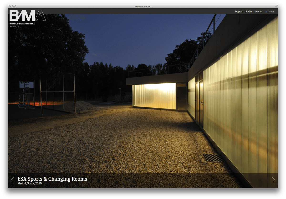 A modern building with illuminated glass walls at dusk, designed by BernuzBenitez Martinez Arquitectos. The structure is surrounded by gravel and a fenced sports area. The text overlay reads "ESA Sports & Changing Rooms, Madrid, Spain, 2020.